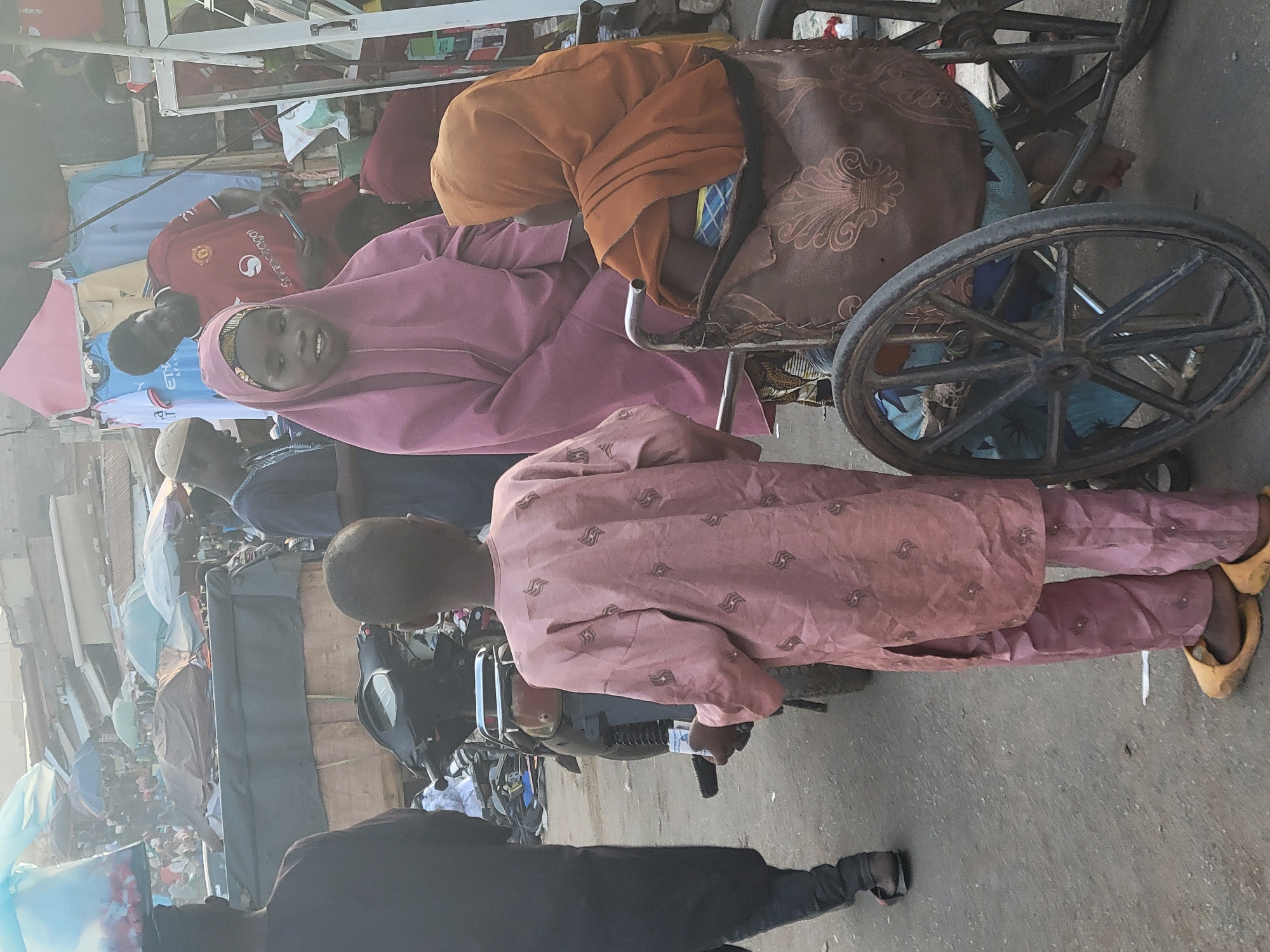 In Madalla, Nigeria, a street scene depicts a person in a wheelchair. A young woman in a pink hijab looks at the viewer. An individual in a pink patterned garment stands next to the wheelchair, near parked motorcycles. Other people are nearby. Clothes are hung in the background. The ground is paved.