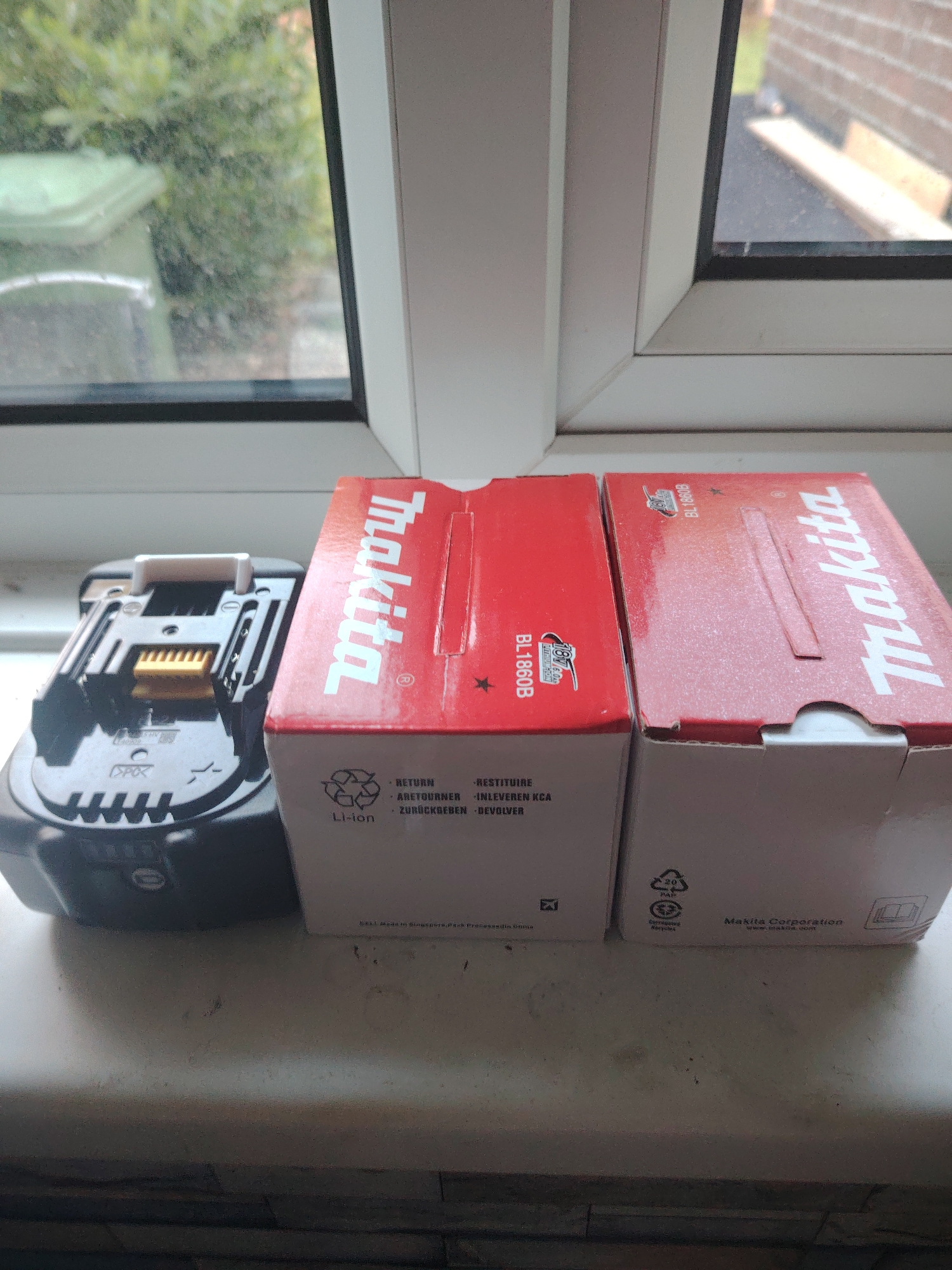 Two red and white Makita battery boxes, labeled "BL1860B," sit on a windowsill. To the left of the boxes is a black Makita battery pack. The windowsill is set against a window with a view of green foliage and a building. The location is the city of Huntingdonshire, United Kingdom.