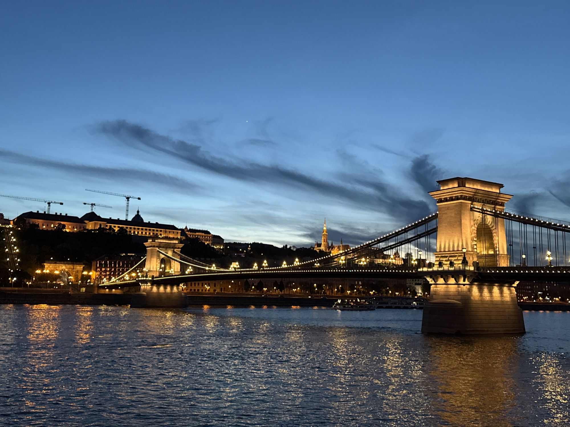 The image captures a vibrant night scene of Budapest, Hungary, showcasing the iconic Széchenyi Chain Bridge (Lánchíd) spanning the Danube River at dusk or early night. The scene is illuminated by numerous warm, golden lights from the bridge and surrounding buildings, creating a striking contrast with the deep blue sky and cool tones of the water.
In the foreground, the Danube River flows, its surface reflecting the city lights in shimmering, elongated streaks due to the gentle ripples. The Chain Bridge is the central subject, with its ornate stone pillars and suspension cables brightly lit. Lights line the bridge deck, forming a bright path across the river.
On the left bank (Buda side), the illuminated buildings of the Buda Castle complex perch atop a hill, with more city lights twinkling below. Several tall construction cranes are visible against the skyline on this side, indicating ongoing development. On the right bank (Pest side), further buildings are visible, including what appears to be the spire of a church or basilica, likely St. Stephen's Basilica or Matthias Church, also lit. At least one boat or river cruise vessel can be seen traversing the water beneath the bridge, also casting reflections.
The sky is a prominent feature, a rich indigo blue transitioning to darker tones. Distinct, wispy, and elongated dark clouds stretch across the sky, creating dramatic patterns. A single, bright point of light, likely a star or planet (possibly Venus), is visible in the upper-middle portion of the sky. The overall impression is one of a bustling, historical city beautifully lit under a twilight sky with interesting cloud formations. There is no visible text or identifiable people.