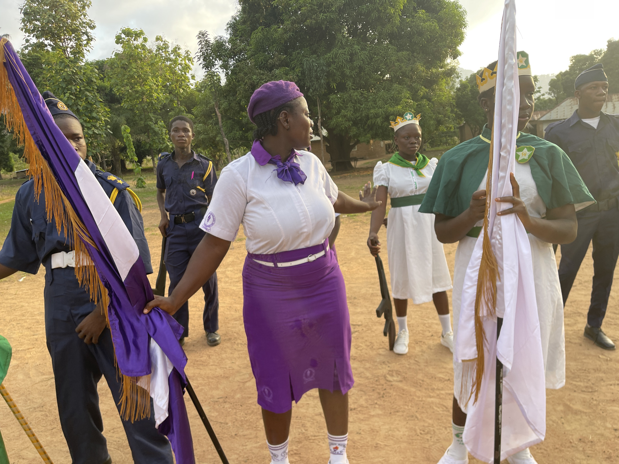 The image depicts a group of individuals outdoors in what appears to be a dirt field, possibly in the city of Andaha, Nigeria. The setting is somewhat rural, with trees visible in the background.
Several people are visible in distinct uniforms. One woman wears a white collared shirt, a purple skirt, and a matching beret. Two individuals are dressed in white garments with green accents. Two males are dressed in dark blue uniforms with hats, and the second has a purple marker in his pocket. Two individuals are holding flags. One flag is a combination of purple and white with gold tassels, and the other is white.
The subjects are interacting, and there appears to be an element of formality or ceremony. The overall atmosphere is one of a gathering or event, with the uniforms and flags suggesting an organizational or institutional context.
