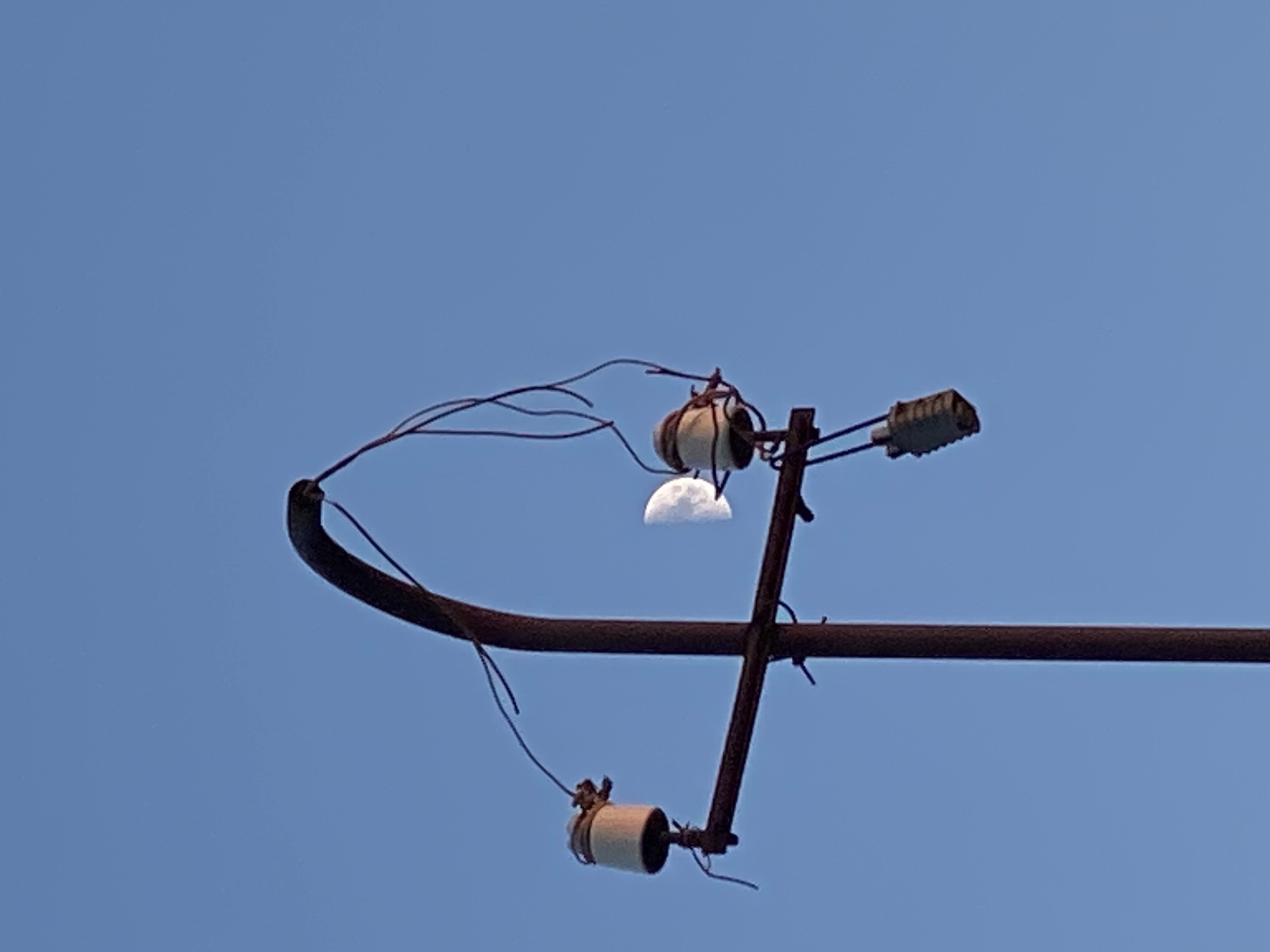 The image depicts a utility pole against a clear blue sky. The pole is made of weathered, reddish-brown metal and has a curved top. Attached to the pole are several electrical insulators, which are off-white ceramic cylinders. Some of these insulators have wires attached to them, appearing to be either intact or broken and dangling. A partial view of the moon, illuminated on its right side, is visible between two of the insulators, appearing to be positioned behind the pole. One insulator has a darker, rectangular object suspended below it. The scene is set in Cairaclia, Moldova.
