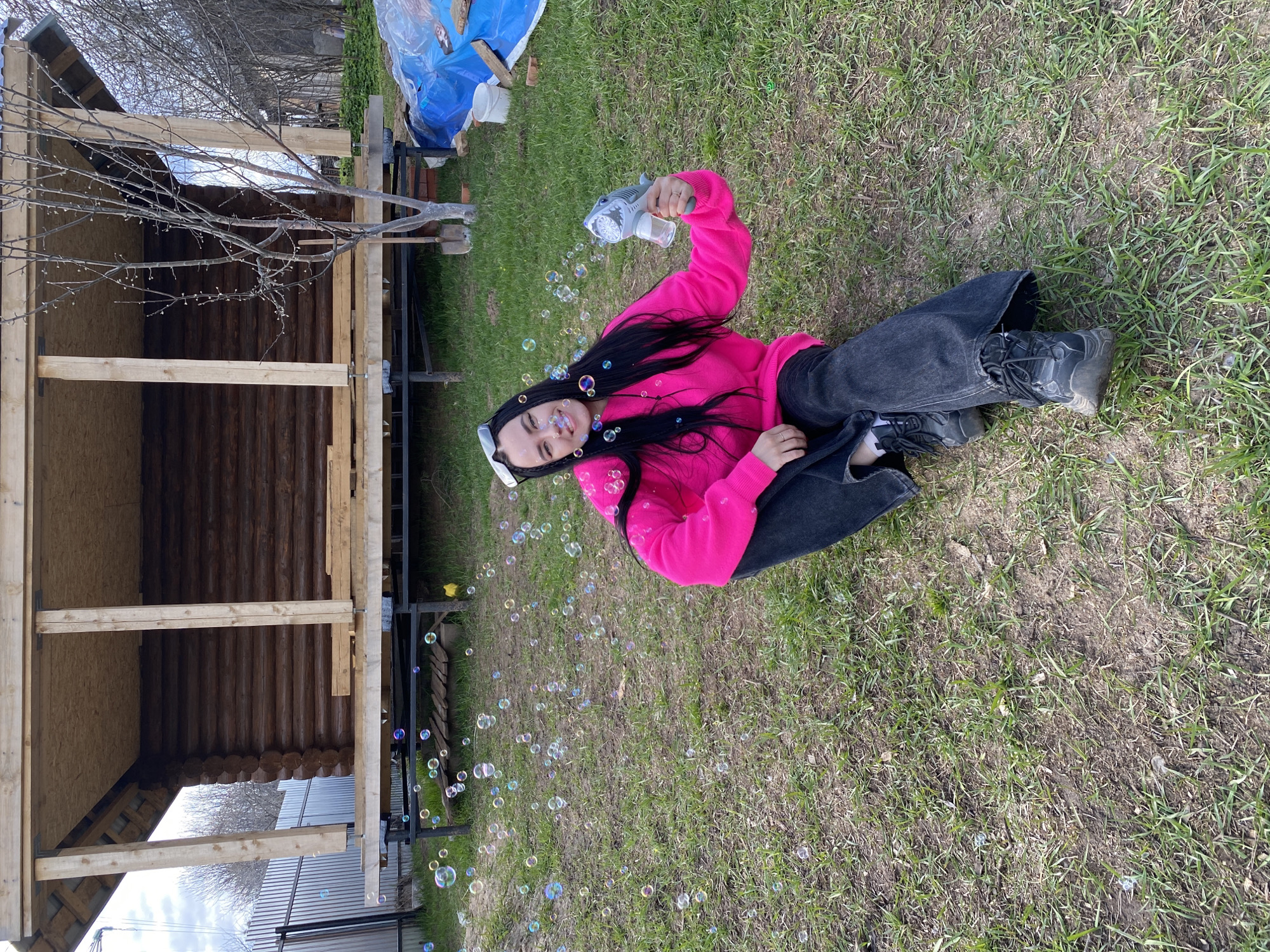 This image depicts a joyful outdoor scene in Chaikovsky, Russia, likely during early spring. The central subject is a young woman with long, dark hair, lying relaxed on a patch of green grass. She is wearing a bright pink long-sleeved top, dark jeans, and dark, chunky shoes. Her sunglasses are pushed up on her head, and small, colorful clips are visible in her hair.
The woman holds a grey, shark-shaped bubble gun in her right hand, from which numerous iridescent soap bubbles are actively being released and are floating around her and across the grassy area. She is smiling broadly and looking towards the camera, conveying a sense of happiness and playfulness.
In the background, to the left, a wooden structure, possibly a gazebo or shed under construction, is visible with exposed timber and log walls. Adjacent to this structure stands a bare tree with numerous small buds, indicative of early spring. Further behind, a blue tarp covers some items, and other construction materials, including bricks and a shovel, are partially visible. The ground is a mix of green grass and patches of soil. The scene is brightly lit, suggesting clear daytime weather. No text is visible in the media file.