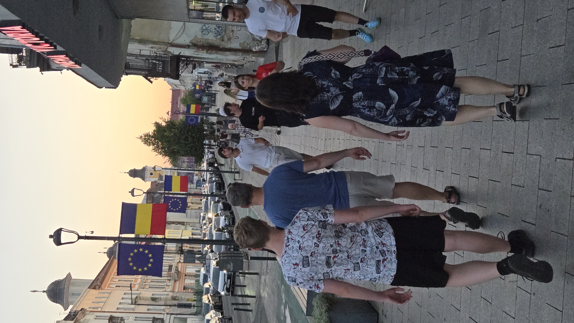 The image captures a street scene in Cluj-Napoca, Romania, with a group of people walking on a paved sidewalk. The time appears to be late afternoon or early evening, given the warm, soft light and the sky's hue. The buildings lining the street are a mix of architectural styles, with one prominent building on the right displaying the illuminated sign "CINEMA VICTORIA." EU and Romanian flags are visible, hung from lampposts, suggesting a public or celebratory event. The street is lined with parked cars on the left. In the foreground, four individuals are walking away from the camera. The person on the far left wears a white t-shirt with a pattern of cartoon characters and black shorts. Next to them, a person in a blue t-shirt and light-colored shorts walks. A woman in a dark blue floral dress is positioned further back on the right, followed by another woman whose face is not visible. Several other people are seen walking in the background, suggesting a moderately busy urban environment. The ground is paved with rectangular stones.