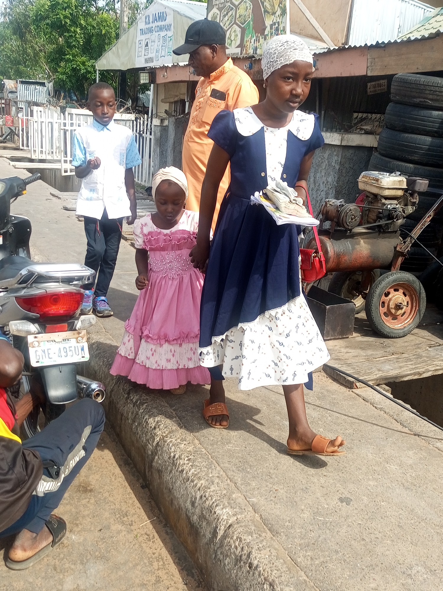 The image captures a street scene in Tempure, Nigeria, with several individuals present. In the foreground, a young girl in a pink dress stands on a concrete curb, while a teenage girl in a blue and white dress walks beside her. The teenage girl holds a bundle of papers and a red purse. Behind them, a young boy in a white and blue shirt and black pants walks with his hands in his pockets. A man wearing an orange shirt and a black cap stands to the right. A motorcycle is parked on the left side of the frame, and in the background, a stall with the sign "KB. JAMI TRADING COMPANY" is visible. Various objects, including stacks of tires and a generator, are present in the environment. The scene appears to be an outdoor market or commercial area, typical of a Nigerian city.