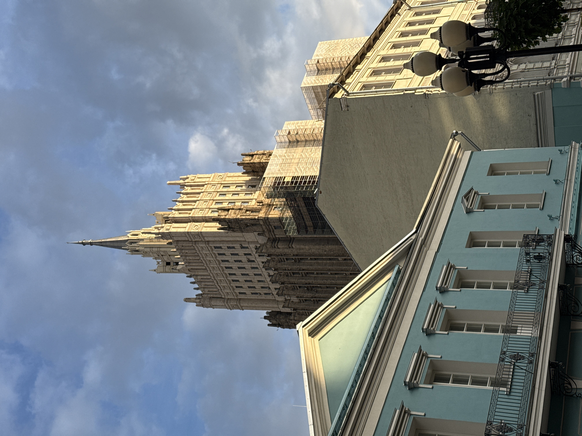 The image presents a striking urban architectural scene in Moscow, Russia, viewed from a low angle looking upwards, creating a sense of grandeur and scale. The overall lighting suggests late afternoon or early morning, with sunlight illuminating the buildings and creating strong shadows under a partly cloudy sky.
The central and most prominent subject is a towering, ornate Stalinist-era skyscraper, likely one of the "Seven Sisters." This multi-storied building is characterized by its intricate, light-colored facade, numerous windows, and a distinctive spire topping its central section. Scaffolding is visible on various parts of the skyscraper, indicating ongoing construction or renovation work.
In the foreground and midground, several other buildings frame the view. To the immediate right, a light blue building with white window frames and elaborate black metal railings on its balconies or sills stands out. Behind it, a plainer, light beige building is partially visible. Further to the right, another multi-story, light-colored building with multiple windows is seen. An ornate black street lamp with several globes extends into the upper right portion of the frame.
The sky is a mix of bright blue and scattered grey and white clouds, providing a dynamic backdrop to the impressive architecture. No human figures or discernible activity are visible, lending a static yet majestic quality to the urban landscape. There is no visible text in the image.