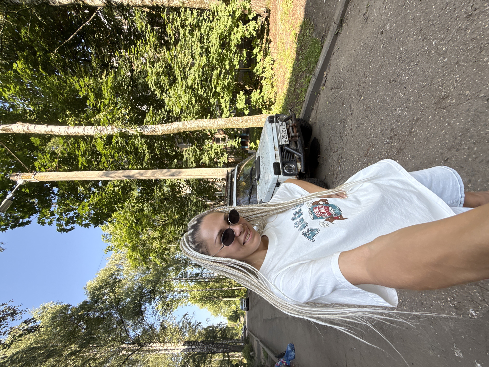 In the city of Rusanovo, Russia, a woman with long, blonde braided hair and sunglasses is taking a selfie on a paved street lined with trees. She is wearing a white t-shirt with a graphic print and light-colored shorts. Behind her, a white off-road vehicle with a roof rack and a modified bumper is parked on the side of the street. The environment is a residential area with trees and buildings visible in the background. The lighting suggests it is daytime, likely late afternoon due to the warm, soft sunlight.