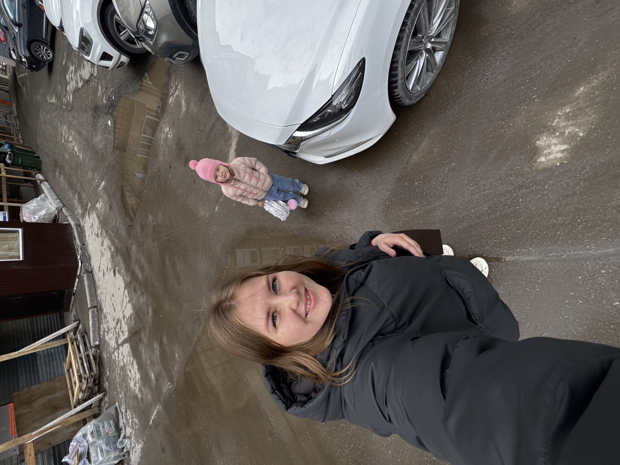 The image captures a woman taking a selfie in an urban environment. A small child, wearing a pink hat and jacket, stands near a white car, facing away from the camera. Several cars are parked nearby on a wet paved surface. The background includes reflections in puddles, wooden pallets, and building facades, suggesting a street or alleyway in Perm, Russia.