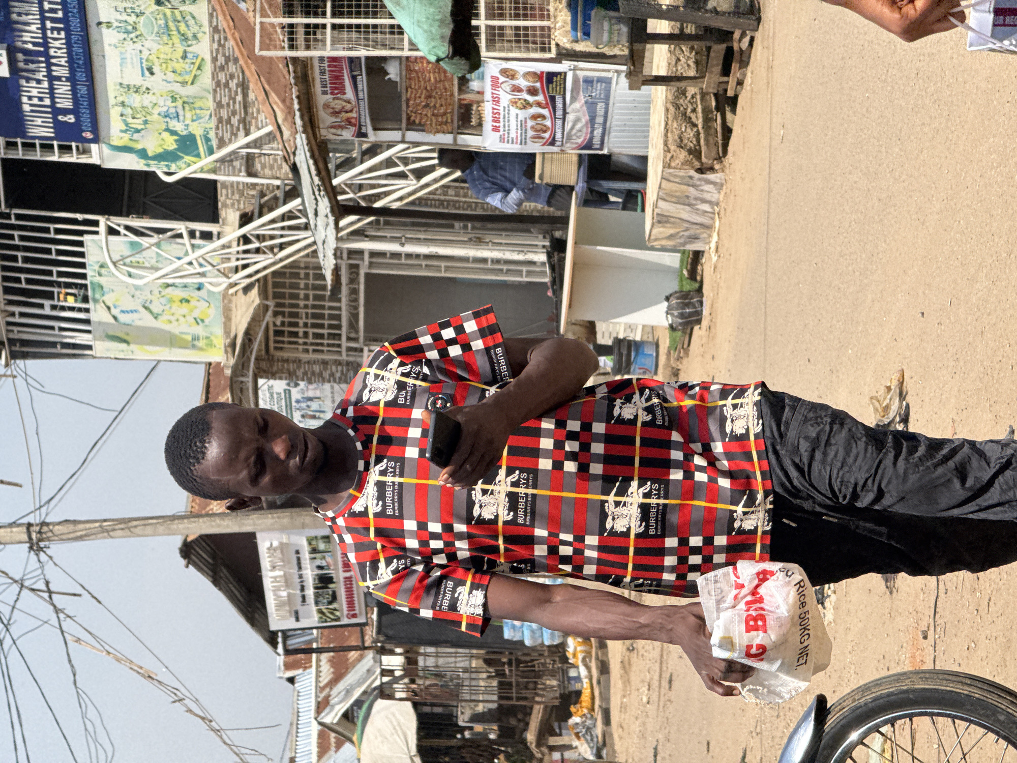 A young Black man walks along a dusty street in Abuja, Nigeria, looking down at his smartphone. He is wearing a red, black, and white plaid t-shirt with the Burberry logo repeated throughout, and dark, possibly black, pants. In his left hand, he carries a white plastic bag with red lettering that reads "RICE 50KG NET." To his right, a motorcycle wheel is partially visible. In the background, there are several market stalls, shops, and residential buildings, with electrical wires crisscrossing overhead. One building in the background has signs for "WHITEHEART PHARMA & MINI-MARKET LTD." and advertisements for fast food.