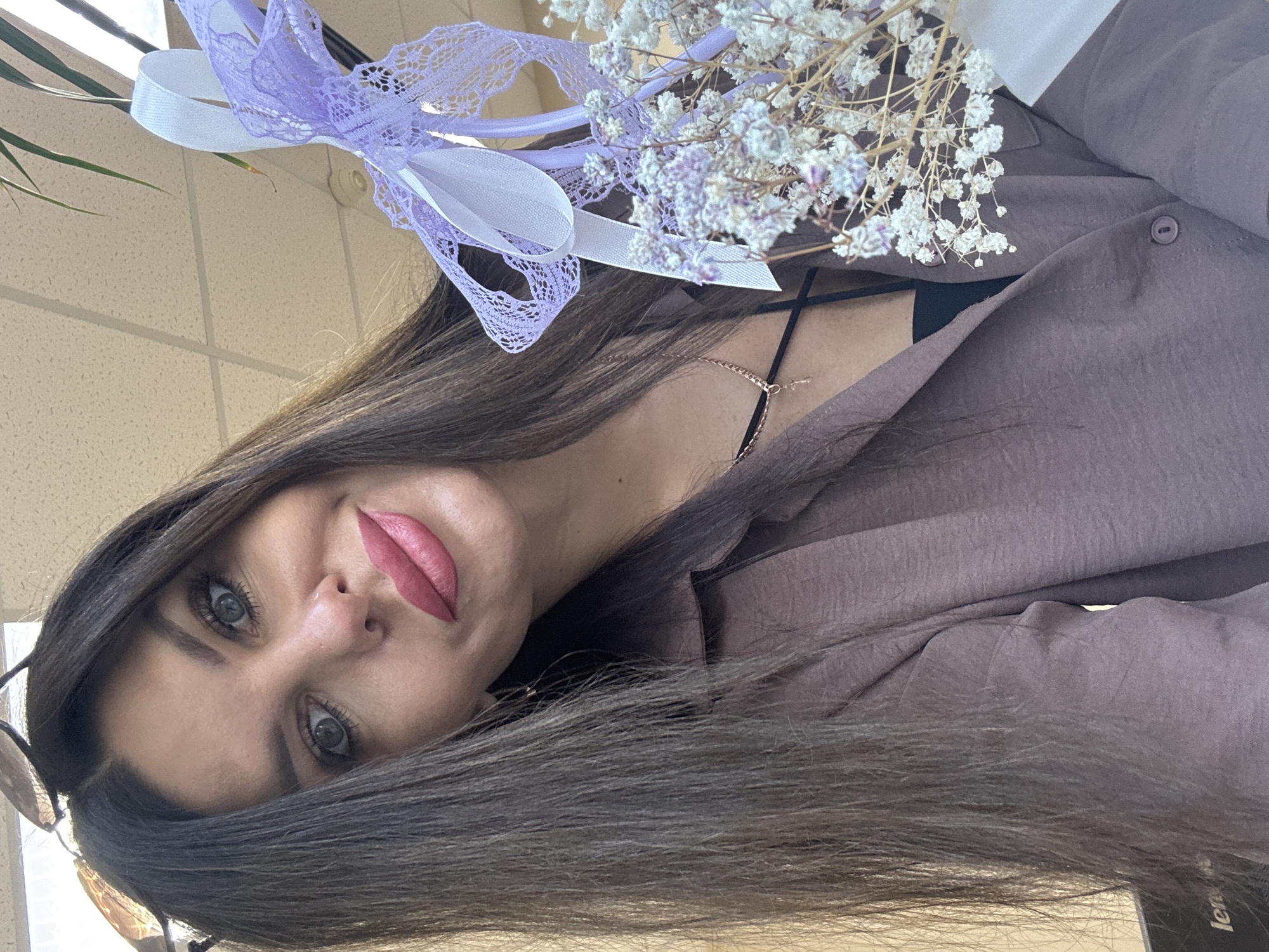A woman with long dark hair and blue eyes is holding a small bouquet of white flowers tied with light purple ribbons. She is wearing a mauve-colored button-down shirt over a black top and a delicate gold necklace with a cross pendant. The background appears to be an indoor office environment with a tiled ceiling and fluorescent lighting. A plant with long green leaves is visible in the upper right corner. Part of a laptop or computer monitor with the word "len" visible is in the lower left corner. The woman is looking at the camera and smiling slightly. The image was taken in Chaikovsky, Russia.