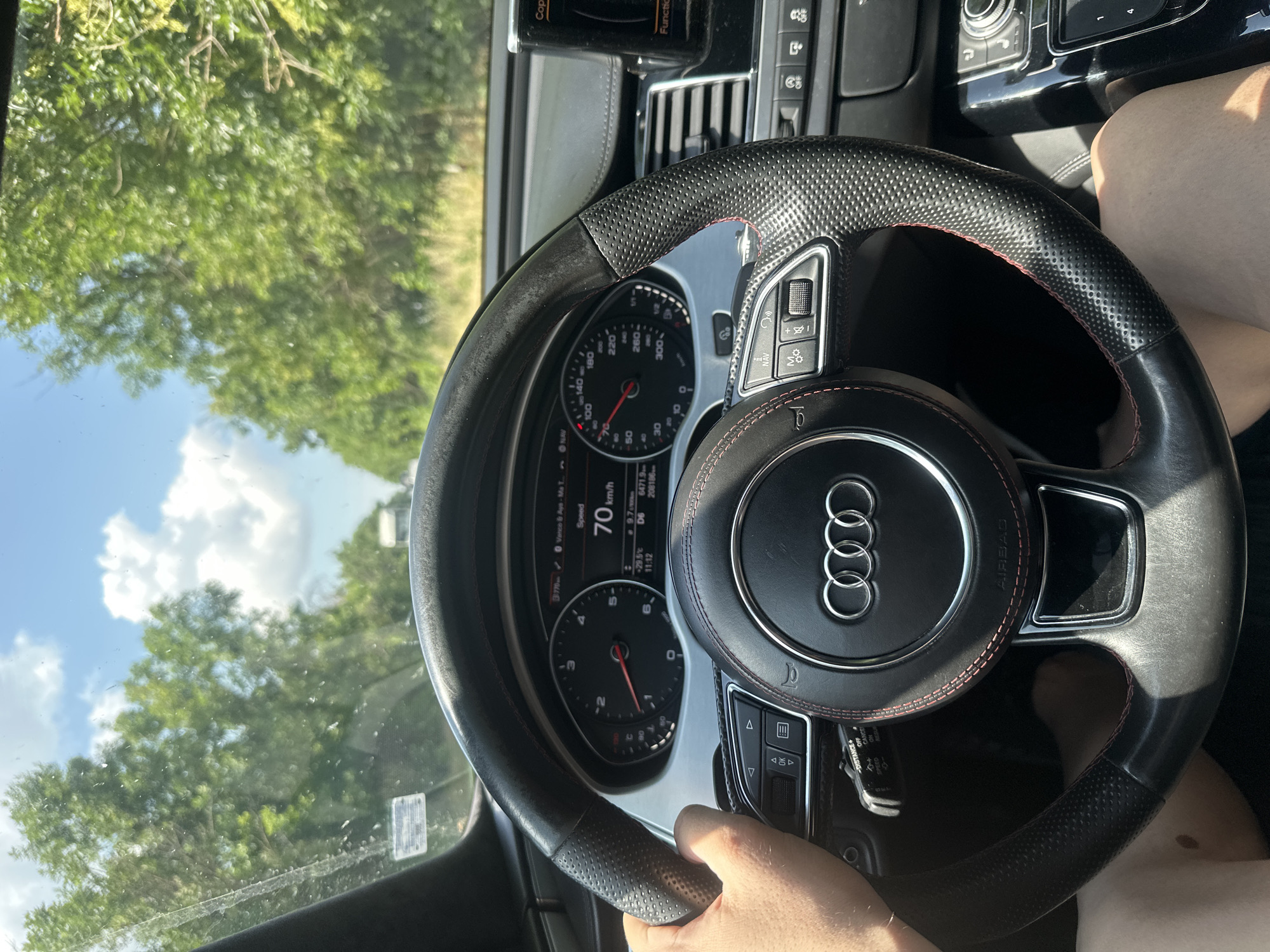 The image shows the interior of a car, focusing on the steering wheel and dashboard, as seen from the driver's perspective. The driver's left hand is visible on the steering wheel. The car is moving, indicated by the speedometer displaying "70 km/h" and the passing scenery of trees and a blue sky with clouds through the windshield. The dashboard features a digital display showing various information including speed, distance, and time. The location is identified as Obretenik, Bulgaria.