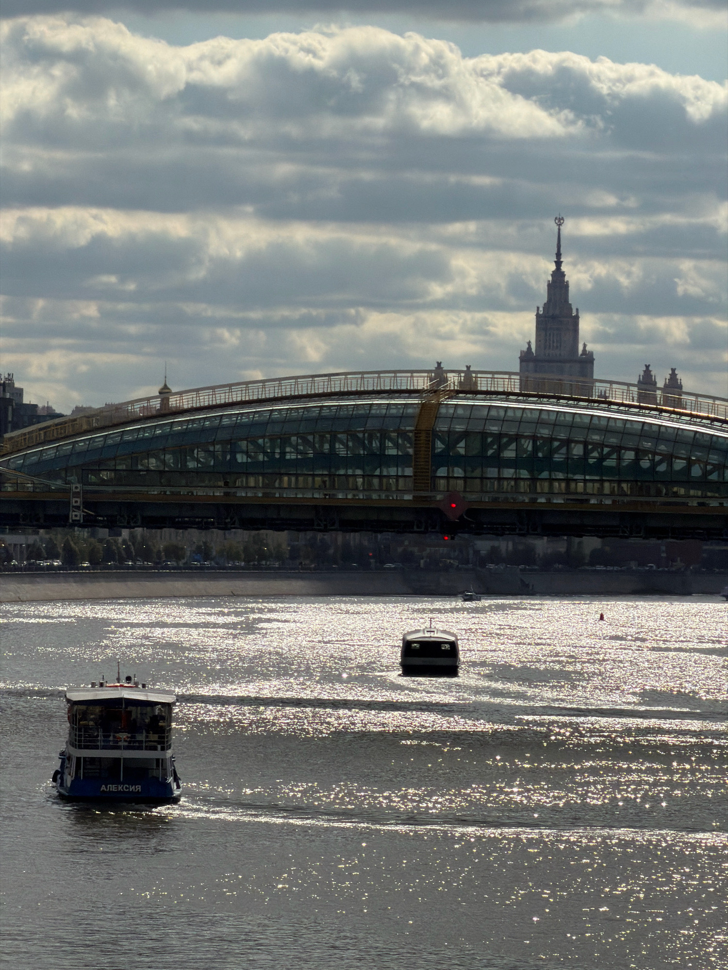 A daytime, medium shot captures a view of the Moskva River in Moscow, Russia, under a dramatic sky filled with heavy, grey clouds interspersed with patches of blue. In the foreground, a blue and white passenger boat, named "АЛЕКСИЯ" in Cyrillic lettering, navigates the water, its occupants visible through the windows. Further in the distance, a smaller, dark vessel is also present on the water. The river's surface is highly reflective, shimmering with the sun's rays. Spanning across the middle ground is a modern bridge with a distinctive arched, glass-covered structure. Behind the bridge, the iconic Stalinist skyscraper, Moscow State University, rises prominently against the cloudy sky, its spire topped with a star. The overall scene conveys a sense of urban activity and dramatic weather conditions.