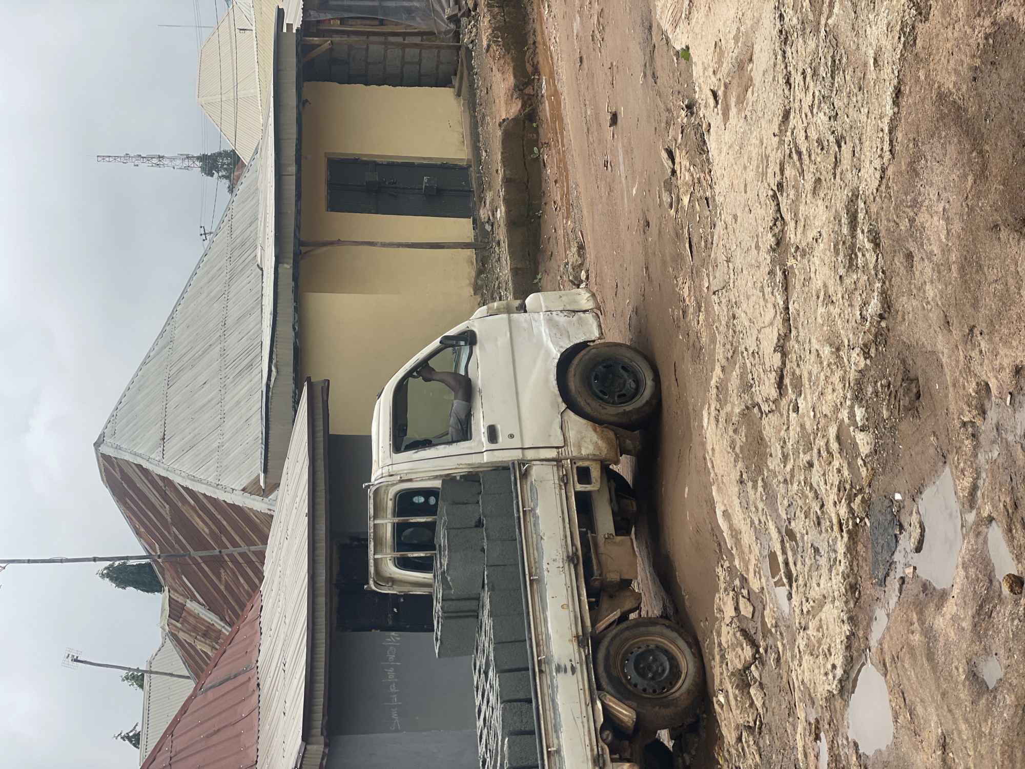 A white pickup truck carrying several stacks of concrete blocks drives on a dirt road in Kurunduma, Nigeria. The truck is moving from left to right, and a person is visible in the driver's seat, with their arm resting on the open window. The road is uneven and appears to have puddles and debris scattered across it. In the background, there are several buildings with corrugated metal roofs and yellow and grey walls. A cell tower is visible in the distance. The sky is overcast.