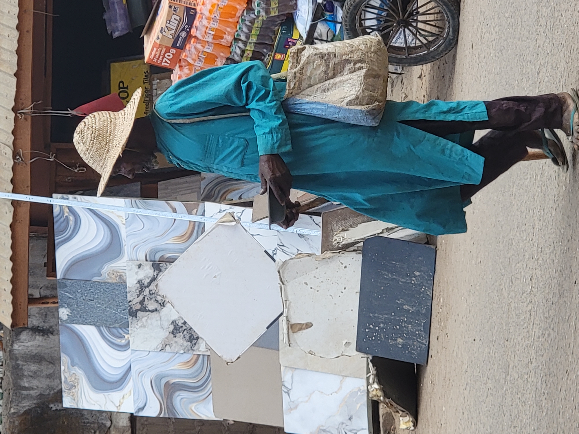 The image depicts a vibrant market scene, likely in Maiburiji, Nigeria, with a focus on a person standing amidst a display of various floor and wall tiles. The setting appears to be an outdoor shop or stall.
In the center, a person is shown wearing a wide-brimmed straw hat, a long-sleeved, teal-colored tunic or gown, and dark trousers. They are also wearing light-colored flip-flops. A beige, possibly fabric, bag is slung over their shoulder. The person's right hand is visible, holding a dark rectangular object, which appears to be a smartphone.
The foreground and midground are dominated by an assortment of decorative tiles. These include patterned tiles with swirling designs in shades of gray, white, and gold, as well as plain white square tiles and darker, possibly black or dark gray, tiles. Some tiles show signs of wear or damage.
In the background, shelves are visible, stocked with numerous packaged goods, including many items in orange packaging. A brown cardboard box with text is visible, and part of a bicycle or motorcycle wheel can be seen in the upper right. Wooden posts or beams suggest the structure of a market stall on the left. The ground is a light-colored, possibly paved or packed earth, surface.
The bright lighting suggests it is daytime under clear weather conditions. The atmosphere is indicative of a busy commercial area.
Visible text includes "Klin", "170g", and "24 sachets" on a brown box in the background. A partially visible yellow sign reads "COP" and "Free Floor Tiles," indicating the sale of tiles at the location.