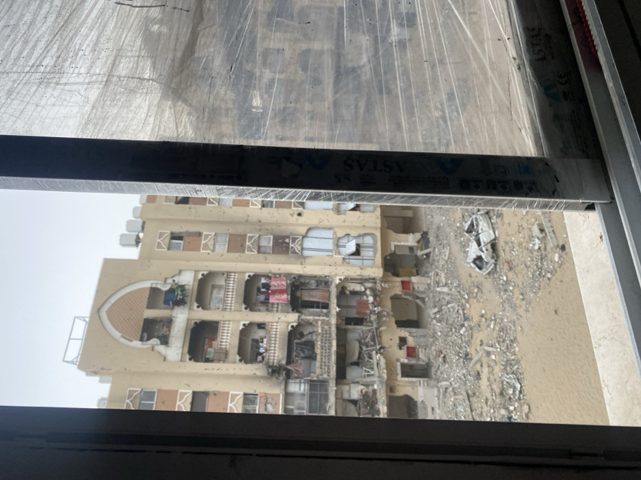 The image depicts a heavily damaged urban scene in Khan Yunis, Palestinian Territory, viewed from an interior window. The viewpoint looks out onto a multi-story, light-colored residential building with balconies, featuring an arched architectural detail at its upper center. The building exhibits extensive structural damage, particularly on its lower floors, with walls partially collapsed, exposed interiors, and numerous pockmarks on the facade consistent with shrapnel or bullet impacts. Burn marks are visible around the central lower balconies. Debris and rubble are strewn across the sandy ground directly in front of the building. A severely damaged, overturned white vehicle, possibly a sedan, lies amidst the rubble on the ground level. Some laundry, including red and white clothes, remains hanging on balconies on the middle floors of the damaged building. The sky is overcast. The foreground shows a window frame on the left and bottom, with a vertical window mullion on the right; the right window pane is covered with transparent plastic sheeting. No individuals are visible in the scene.