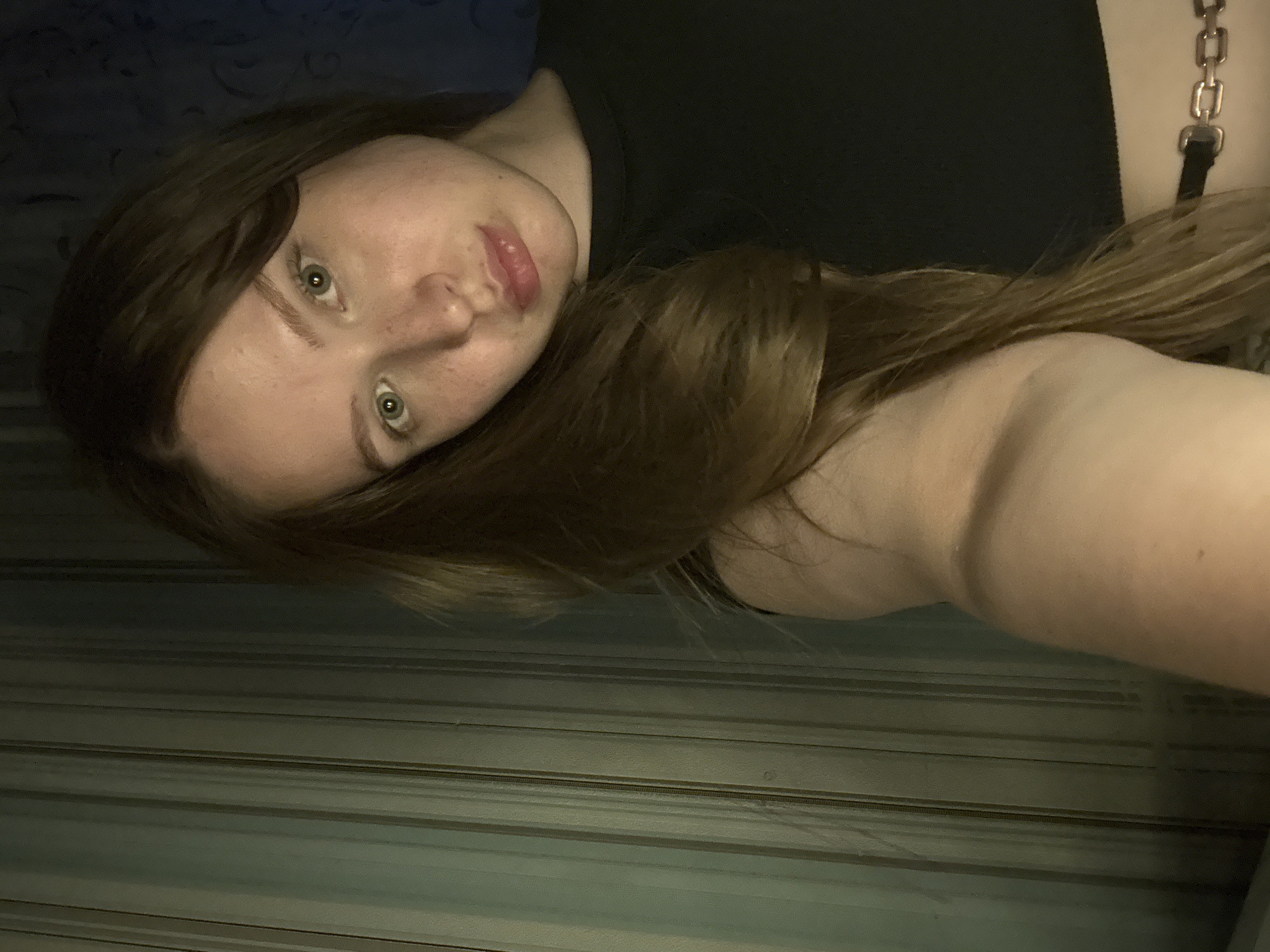 A young woman with light skin, brown hair, and green eyes is lying down and looking at the camera. She is wearing a black top. The background appears to be a wall with horizontal lines and a patterned area, possibly a headboard. There is no information provided in the image to ascertain its location in Chaikovsky, Russia.