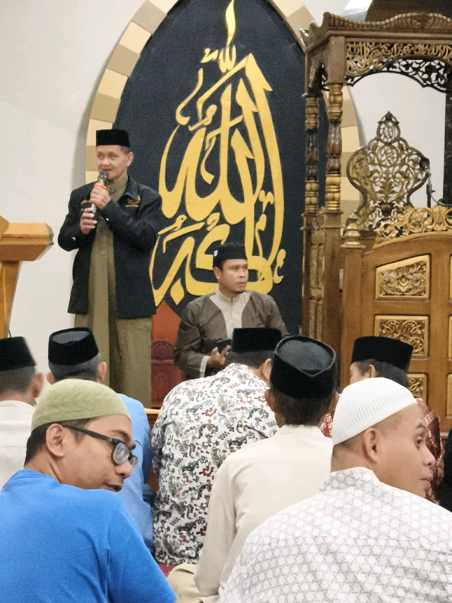 In Makassar, Indonesia, a group of men are gathered inside a mosque. A man wearing a black cap and dark jacket stands at a podium, holding a microphone and speaking. He is dressed in a long, olive-colored robe. Behind him, a large black backdrop features Arabic calligraphy in yellow. To his right, a wooden structure with ornate carvings, possibly a minbar or a decorative screen, is visible. Several men in the foreground, all wearing traditional Islamic caps (peci and kufi), are seated and facing away from the camera, appearing to listen to the speaker. One man in the front left wears a green skullcap and glasses, and a blue t-shirt. Another man in the front right wears a white kufi and a patterned shirt. The overall atmosphere suggests a religious gathering or sermon taking place within a mosque.