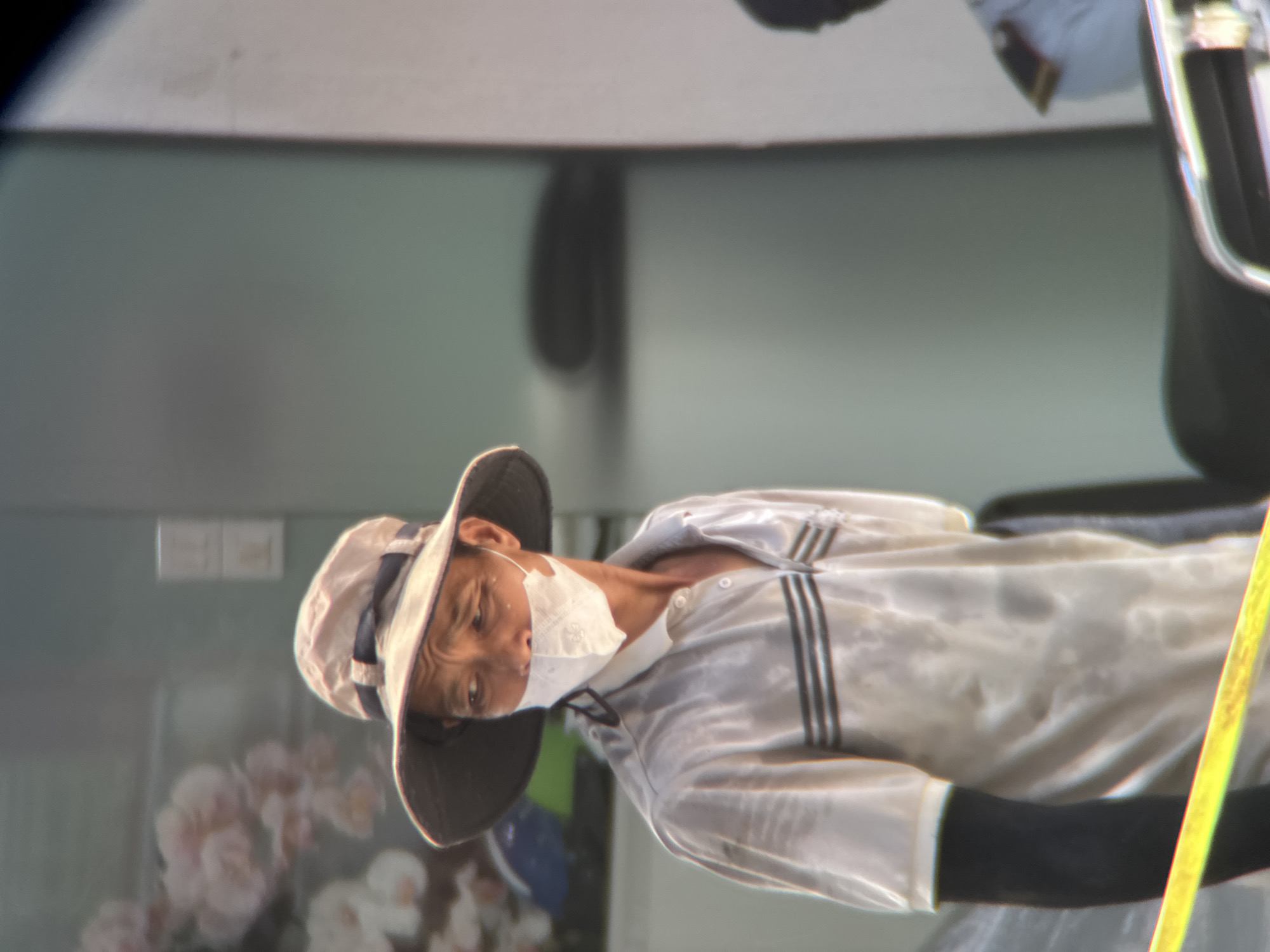 The image shows a person wearing a wide-brimmed hat and a white mask, a light-colored collared shirt with dark stripes, and black arm sleeves. The shirt appears wet with sweat. In the background, there is a wall with floral decorations on the left, and a blurred out section of a building with what appears to be a white wall on the right. There is also a yellow barrier in the foreground. The scene appears to be outdoors, possibly on a street in Da Nang, Vietnam, given the style of dress and the environmental context.