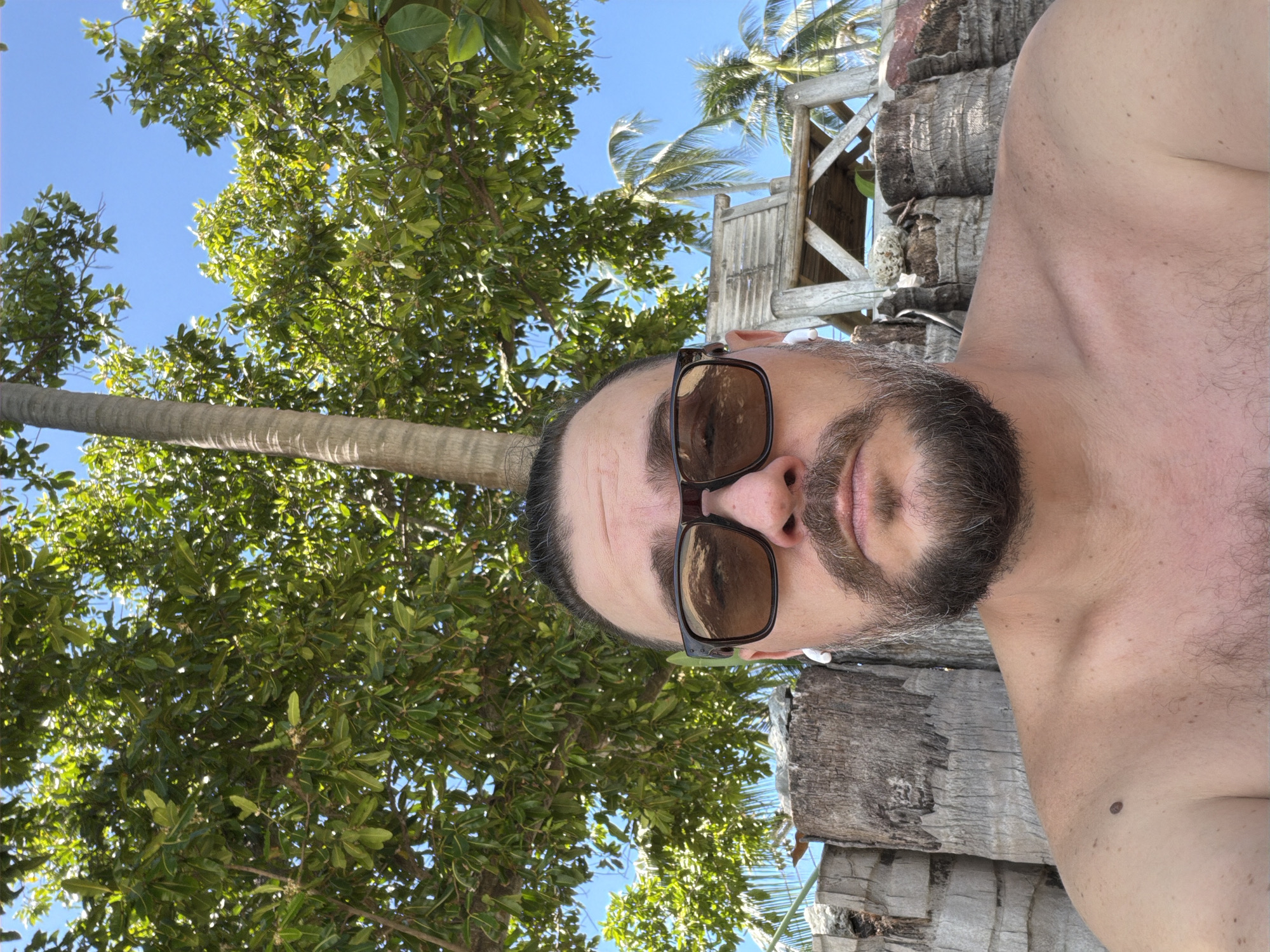 A close-up, over-the-shoulder selfie shows a man with dark hair pulled back, a beard, and sunglasses, wearing white wireless earbuds. He is shirtless, with a mole on his left shoulder. Behind him, palm trees and lush green foliage are visible against a clear blue sky. A wooden lifeguard stand can be seen in the background, along with a rustic wooden fence made of logs. The scene appears to be a beach in Baan Tai, Thailand.
