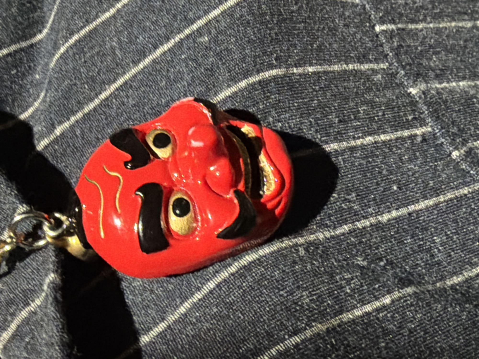A red, mask-like object with black and gold accents, resembling a Japanese Oni mask, is lying on a textured, grey fabric with white pinstripes. A chain is attached to the mask's left side. The scene is lit by direct sunlight, casting a distinct shadow. Location information is unavailable.