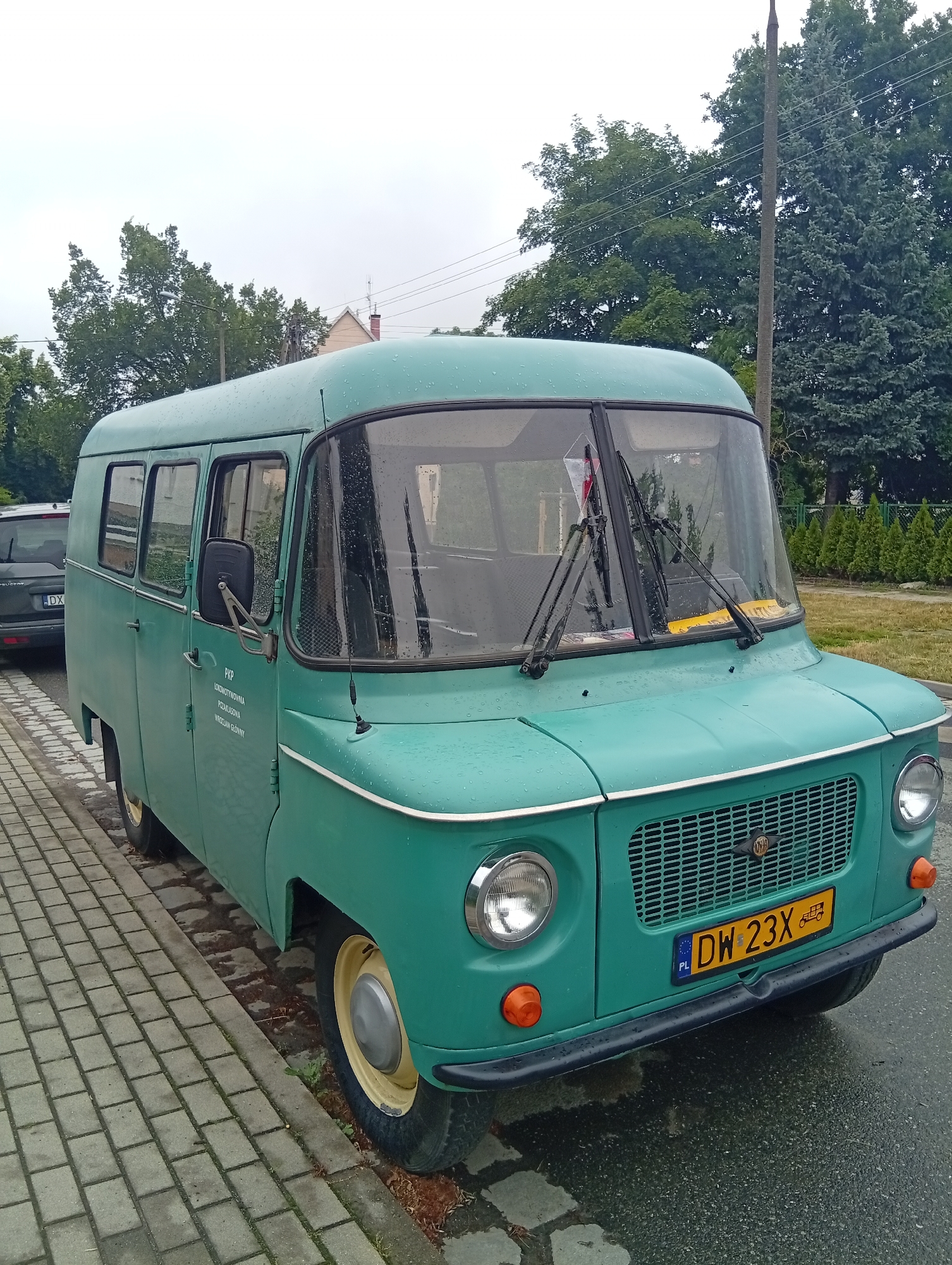 This image shows a vintage, teal-colored van parked on a wet, cobblestone street in Wrocław, Poland. The van appears to be a retro model, possibly from the 1960s or 1970s, with its rounded edges and distinctive grille. It has several windows along its side and a large windshield. The weather is overcast and rainy, as evidenced by the water droplets on the van's surface and the wet road. In the background, there are trees and a power line pole, suggesting an urban or suburban setting. A portion of a dark-colored car is visible to the left of the van.
Visible text on the van includes a license plate with the characters "DW 23X" and the country code "PL". There is also text on the side of the van, appearing to be in Polish, which reads "PKP", "LOKOMOTYWOWNIA", "POZNAŃSKA", and "WROCŁAW GŁÓWNY". The scene suggests a still moment, possibly during a break or before the van is put into use. The overall mood is somewhat muted due to the weather and the vintage nature of the vehicle.