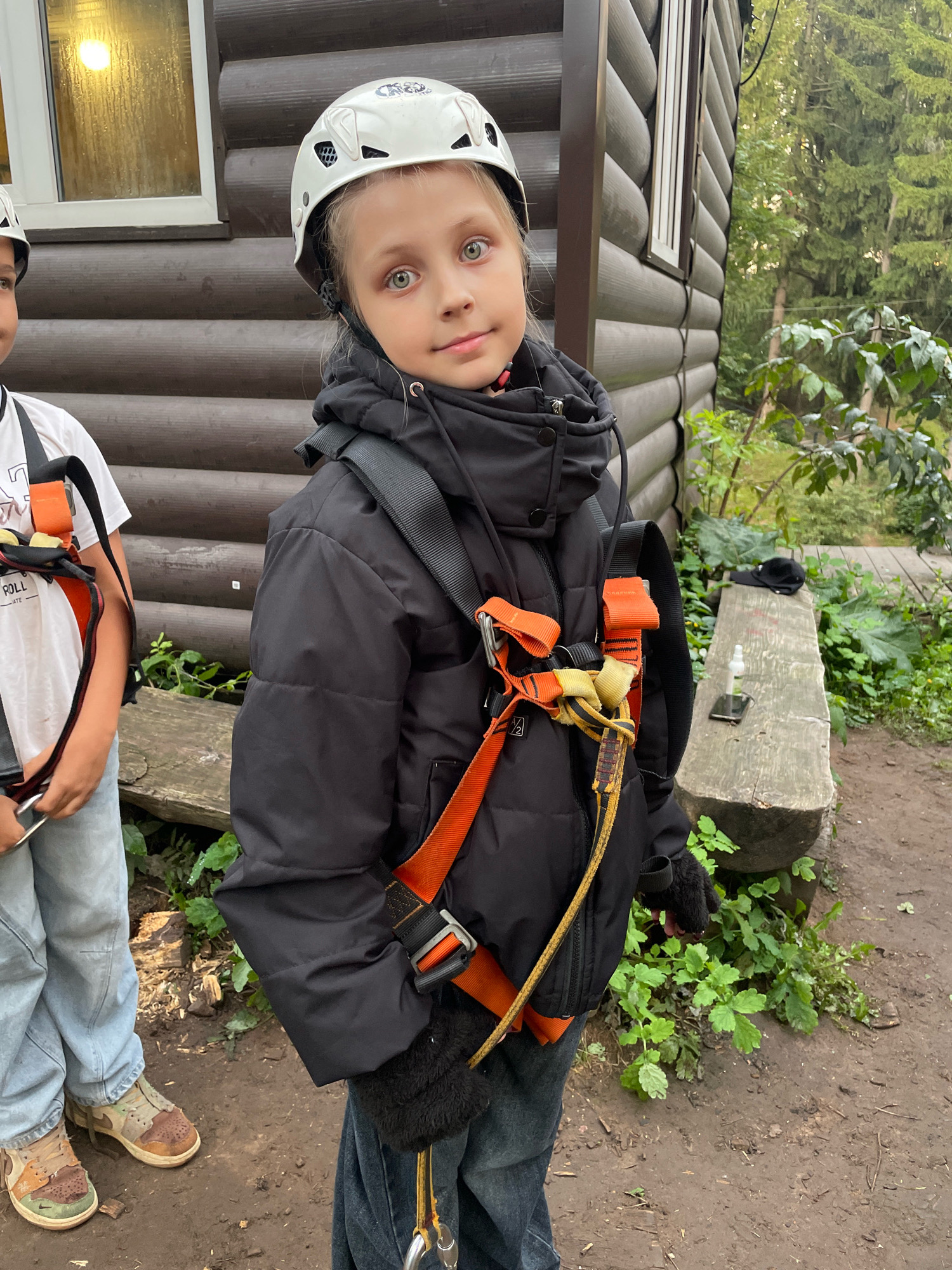 The image shows two children standing outside a wooden cabin in what appears to be an outdoor activity park. The child on the right, a girl with blonde hair and blue eyes, is wearing a white helmet and a black puffer jacket with an orange and yellow safety harness. She is looking towards the camera with a slight smile. The child on the left, partially visible, is wearing a white t-shirt with a graphic print and a similar safety harness. They are both wearing jeans. The background consists of the wooden cabin, lush green foliage, and a dirt path. A wooden bench with a cap, a phone, and a bottle of hand sanitizer is visible to the right. The overall setting suggests an adventurous outdoor activity such as zip-lining or a ropes course. The city of Pervomaysky, Russia, is known for its natural beauty and recreational areas, making it a plausible location for such a scene.
