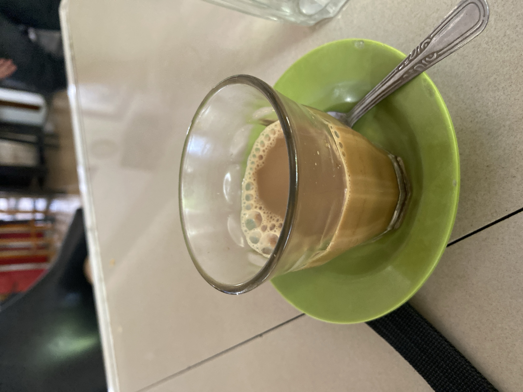 The image displays a close-up, rotated view of a glass of frothy, milky beverage served on a green saucer with a spoon, set on a light-colored table. The scene is likely an indoor setting, possibly a cafe, restaurant, or a traditional eatery in Lhokseumawe, Indonesia.
The main subject is a clear glass filled with a light brown or tan-colored drink, topped with a thick layer of small, uniform bubbles or foam, characteristic of beverages like Teh Tarik or Kopi Tarik popular in the region. The glass is resting within a bright lime-green, circular saucer, which appears to be made of plastic or ceramic. A metallic spoon, likely silver-colored, lies diagonally across the saucer. The spoon has a simple, decorative, embossed pattern along its handle.
The table surface beneath the saucer and glass is light-colored, possibly a beige or light grey, with subtle lines indicating it might be tiled, laminated, or paneled. The background is blurred, showing indistinct dark shapes and a horizontal reddish strip, suggesting other furniture or elements in the establishment. In the lower right corner, a black, textured strap, possibly from a bag or chair, is visible.
No specific activity is captured beyond the presentation of the drink, which appears ready for consumption. The time of day and weather are not discernible. There is no visible text in the image.