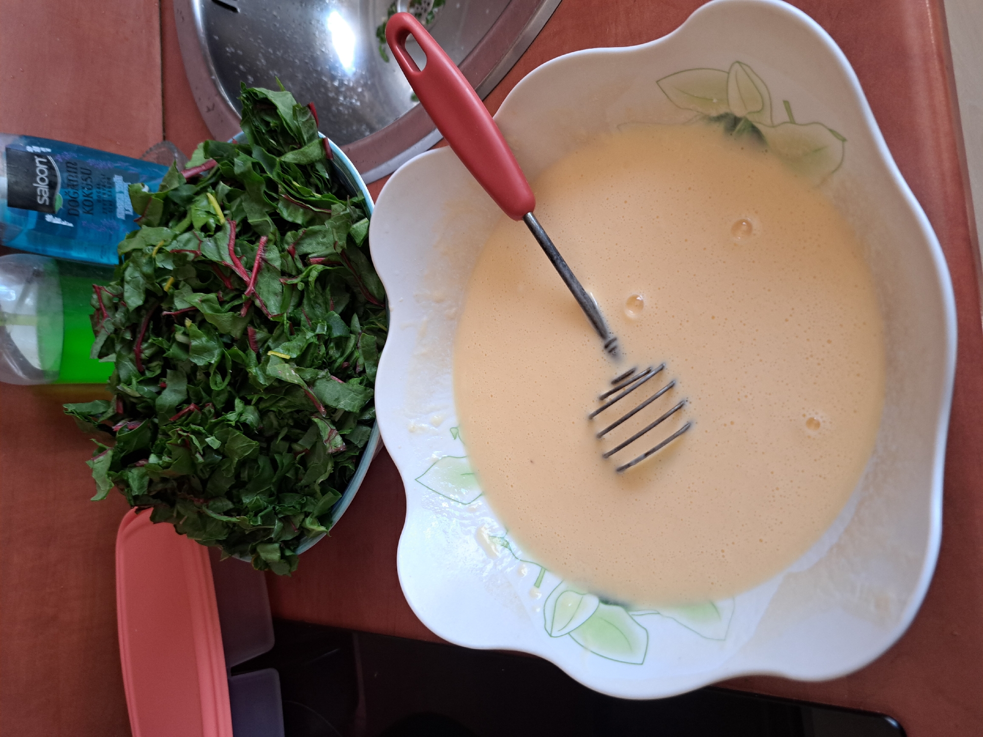 This is a top-down, medium shot of ingredients for cooking. On the left, a blue bottle with the text "saloon" and "DOĞAL KOKUSU" is visible, along with a bowl of chopped green leafy vegetables with red stems. To the right of the vegetables, a white, decorative bowl contains a light yellow liquid, likely a batter, with a red-handled whisk submerged in it. The background shows a metallic surface, possibly a sink, and a brown countertop. The scene appears to be in a kitchen or a food preparation area. The lighting suggests it could be daytime.