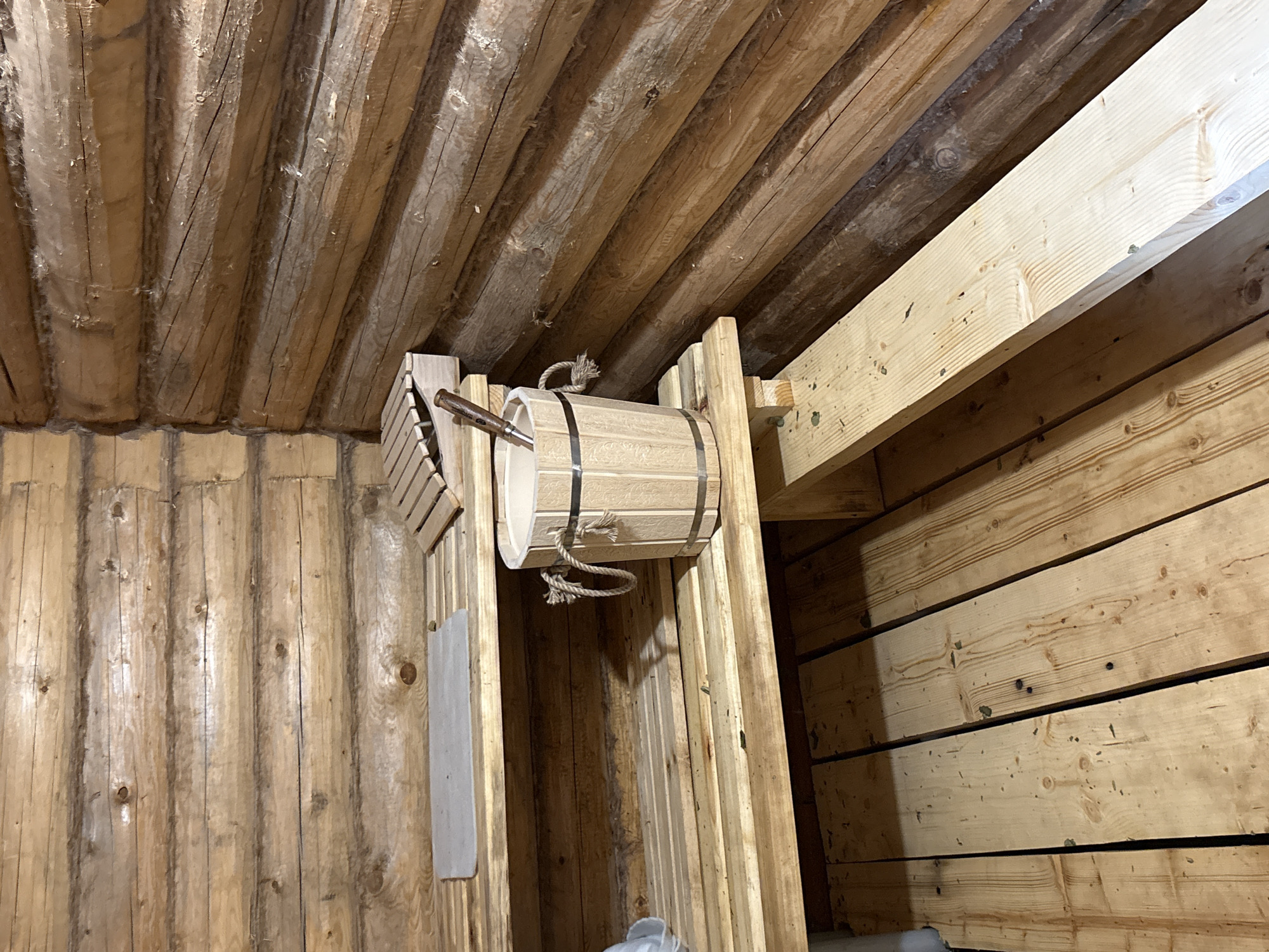 The image shows the interior of a wooden sauna, likely in a private setting. The walls and ceiling are constructed from unfinished planks of wood. A light-colored wooden bucket with metal bands hangs on a vertical wooden support structure. A dipper is placed on the bucket. The lighting is dim, highlighting the texture of the wood.