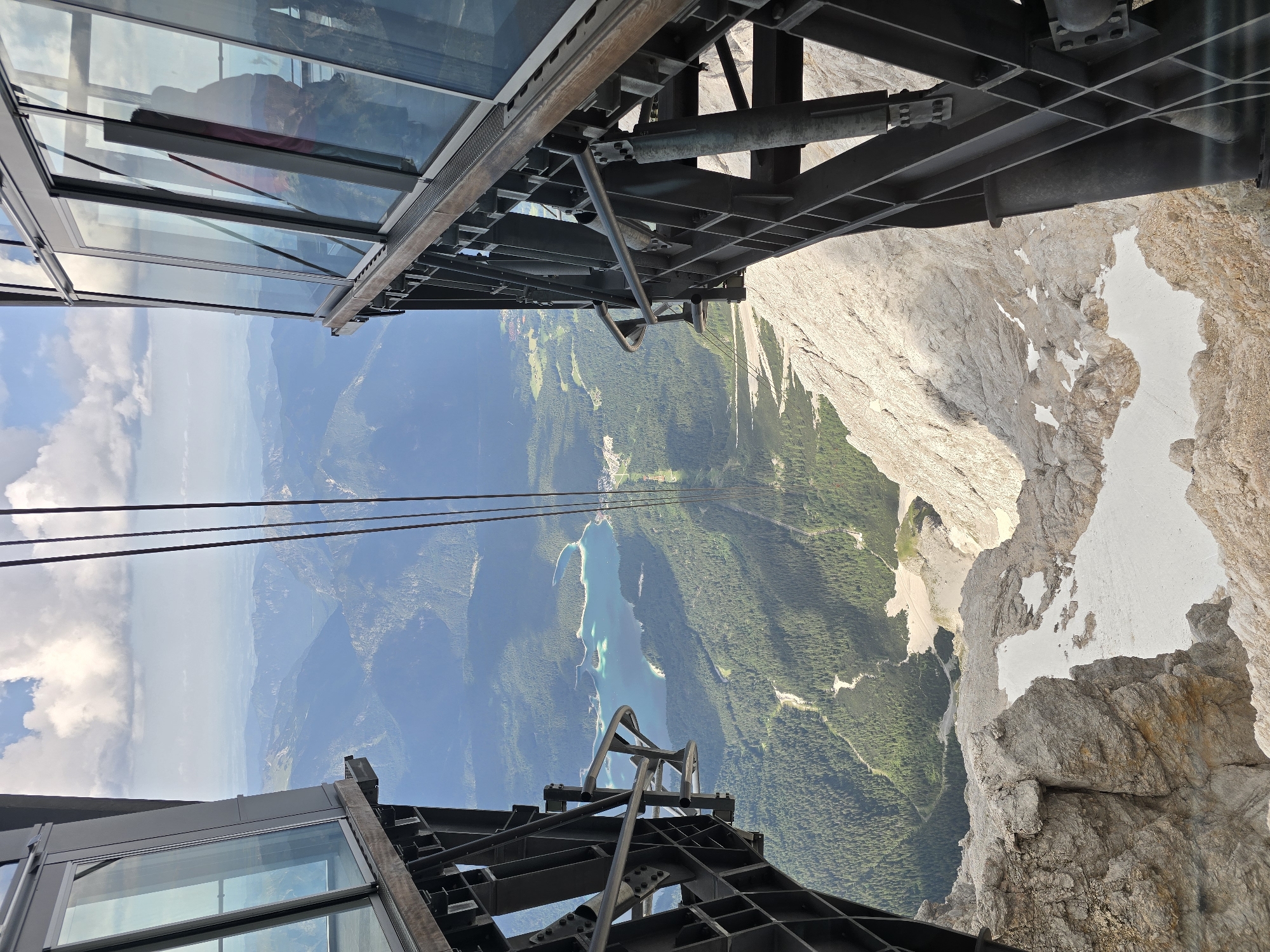 The image captures a breathtaking panoramic view from the top of a mountain, likely the Zugspitze, Germany's highest peak, near Garmisch-Partenkirchen. The viewpoint is from a modern cable car station, with its sleek glass panels and sturdy metal framework dominating the foreground. Below, a vast expanse of rugged, snow-dusted mountainous terrain unfolds, giving way to a lush green valley dotted with pine forests. A striking turquoise lake, Eibsee, is nestled within the valley, its clear waters reflecting the sky. The cable car's thick steel cables stretch tautly towards the horizon, disappearing into the distance. The sky above is a canvas of white, fluffy clouds against a clear blue backdrop, suggesting fair weather. The overall impression is one of awe-inspiring natural beauty and human engineering, offering a dramatic perspective on the Bavarian Alps.