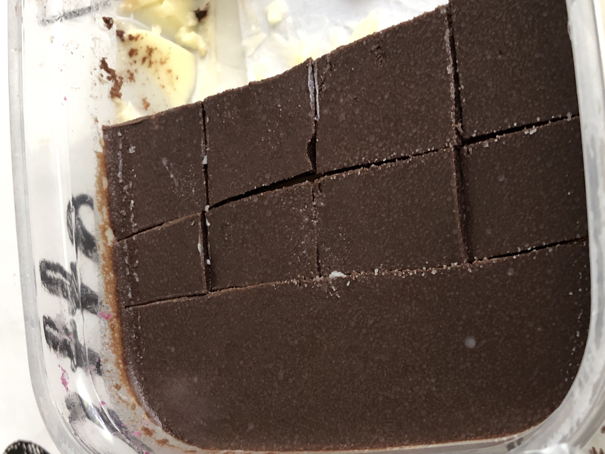 A close-up, overhead shot depicts a block of dark brown, granular-textured food, likely fudge or chocolate, resting inside a clear plastic container. The food has been partially cut into several rectangular portions, with varying depths in the cuts; some are neat lines, while others show more jagged breaks and white residue, possibly sugar bloom or solidified fats, along their edges and on the surface.
In the upper left background, out of focus, a lighter, creamy substance, possibly white chocolate or dairy, is visible with scattered brown crumbs or specks. The clear plastic container shows some wear and tear, with a slight brownish stain or residue along its bottom left interior edge, and a few faint pink specks. Beneath the container on the left, indistinct black markings are visible on the surface. The scene suggests an indoor setting, such as a kitchen, where food has been prepared and portioned. No individuals are present, and the time of day is not discernible.