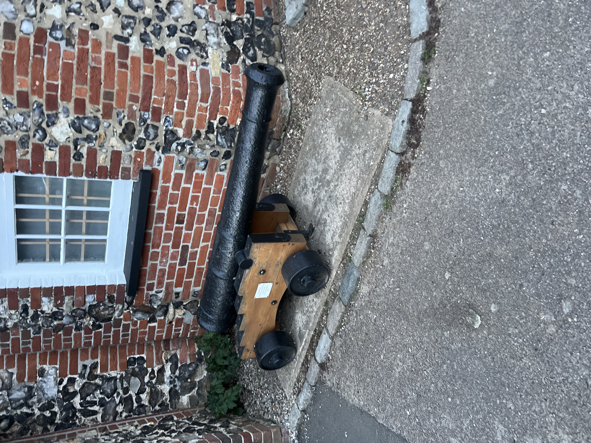 The image depicts a cannon on a wooden carriage positioned beside a brick and flint building in East Suffolk, United Kingdom. The building has a white-framed window and is constructed with alternating layers of red bricks and dark flint stones. The cannon is black and mounted on a light-colored wooden carriage with black wheels. The cannon and carriage are sitting on a concrete surface next to a paved road with a stone curb. Small green plants are visible near the base of the cannon. The image is taken from a high angle.