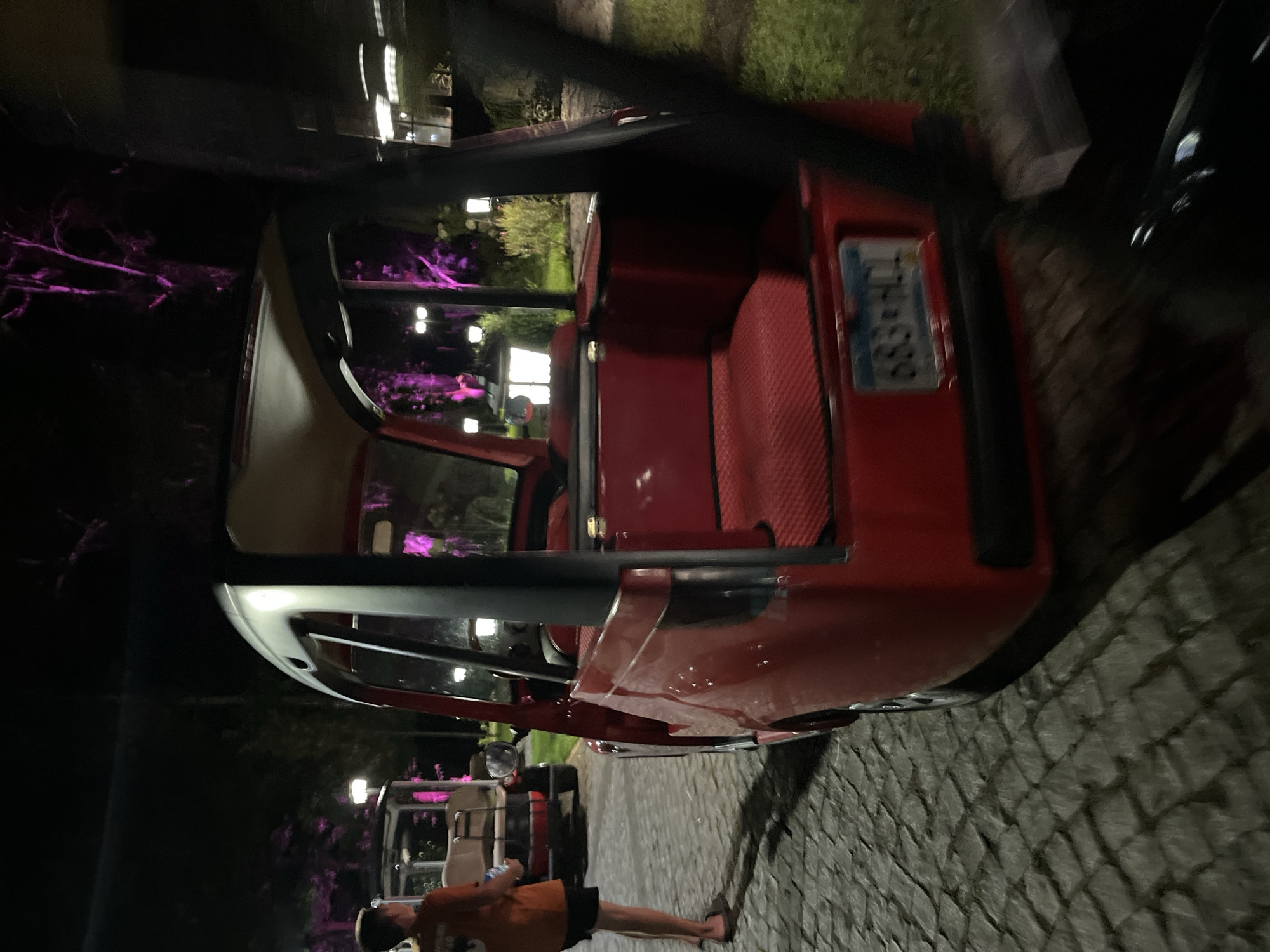 A low-angle, medium shot shows the rear and right side of a red, open-air, multi-passenger vehicle parked on a cobblestone pathway at night in Shekvetili, Georgia. The vehicle has a beige roof and black metal framing. Its interior features red, textured seating and a red floor. A blue and white license plate reading "683-HCL" is affixed to the rear. To the left, a person wearing an orange shirt and black shorts stands facing away from the camera, holding a white object. Another similar vehicle is visible in the background. The environment is illuminated by various light sources, including streetlights and a purple light that casts a glow on trees and foliage, suggesting an outdoor event or park setting. The ground is paved with interlocking cobblestones, and patches of green grass are visible to the right.