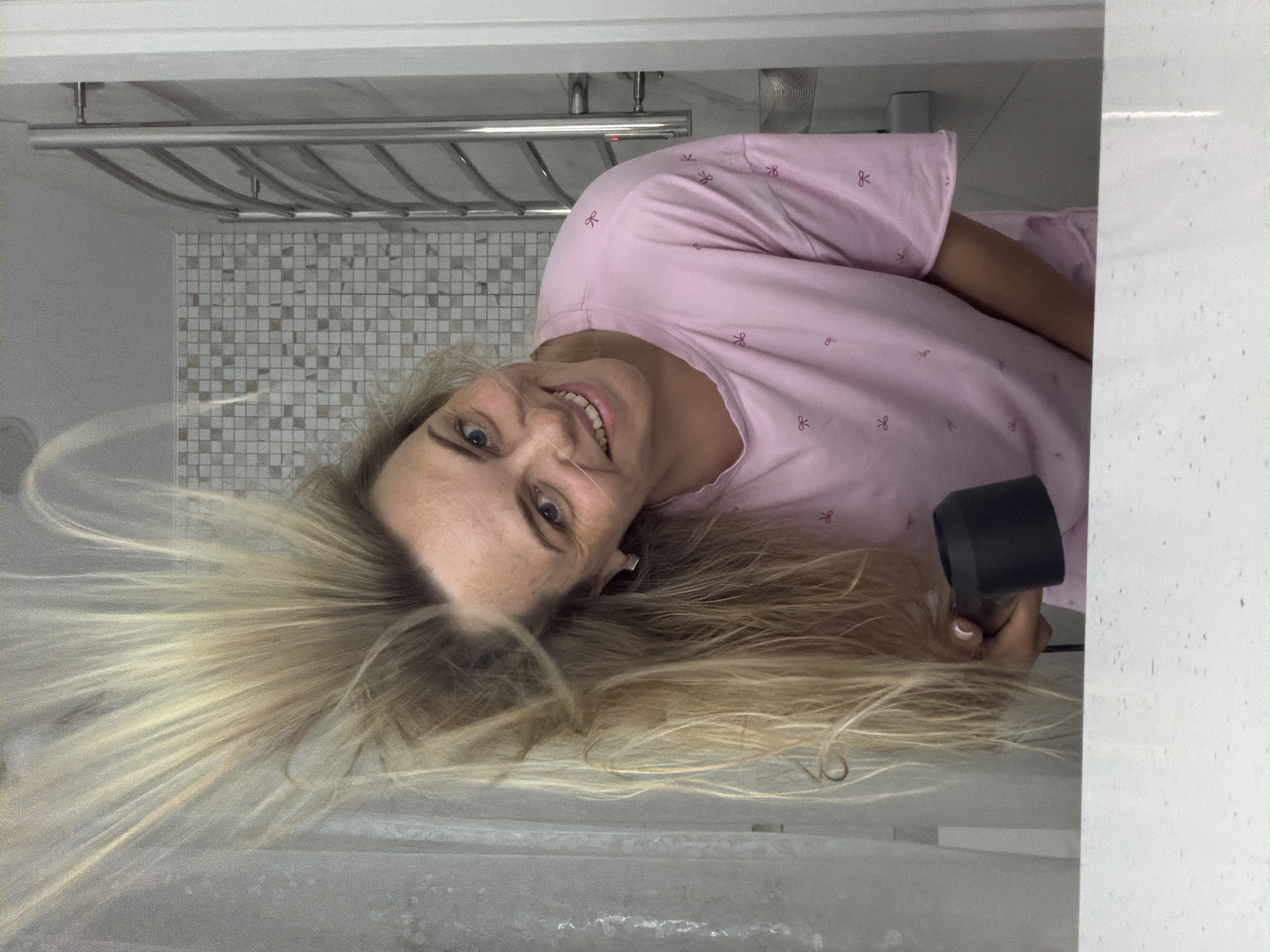 A blonde woman is depicted smiling brightly while drying her hair in a bathroom. The image is rotated 90 degrees clockwise, showing the woman's head and upper torso from a slightly elevated perspective, as if she is lying down and looking up at the camera. She has light skin and blue eyes, and her mouth is open in a wide, cheerful smile, revealing her teeth.
Her long, light blonde hair is dramatically fanned out around her head, appearing to be actively blown by air from a hairdryer. She holds a black, compact hairdryer in her right hand. She is wearing a light pink, short-sleeved top, possibly pajamas or a casual shirt, which is patterned with small, dark pink bow shapes.
In the background, a silver-toned metal towel rack with multiple bars is mounted on a wall. Below the towel rack, the wall is covered with small mosaic tiles, featuring an irregular pattern of white and light gray squares. To the right, a plain white or light gray speckled surface, possibly a wall or vanity, is visible. The scene suggests an indoor, well-lit environment, characteristic of a modern bathroom. The woman's expression conveys happiness and engagement with the activity of drying her hair. No visible text is present in the image.