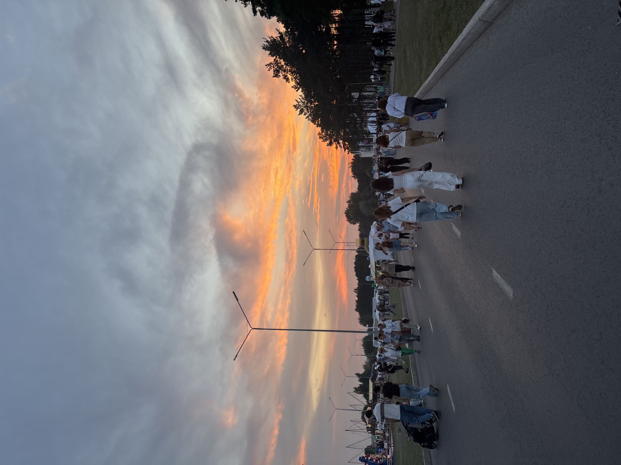 The image depicts a crowd of people walking along an asphalt road during sunset. The sky is filled with vibrant orange and pink clouds, indicating a dramatic sunset. Several lampposts are visible along the road, and trees line the right side of the image. In the distance, there appear to be amusement park rides or stalls, suggesting a festival or fair. People are dressed in casual clothing, and some are pushing strollers or carrying bags. The overall scene suggests an outdoor public gathering or event. It is a photograph taken on the territory of the Central Park of Culture and Recreation in Naberezhnye Chelny, Russia.