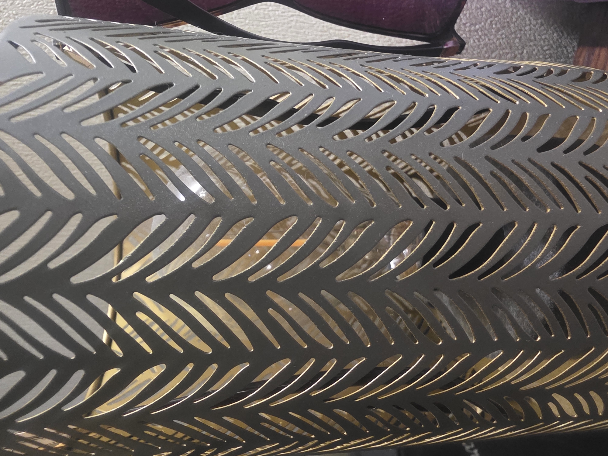 The image shows a close-up of a dark gray metal lantern with a cut-out pattern of diagonal lines that resemble feathers or leaves. The lantern is cylindrical and appears to be hollow, with a warm, golden light emanating from within, suggesting a light source is present. The pattern creates a shadow-like effect on the inner surface. To the right of the lantern, a portion of what appears to be a purple object, possibly sunglasses or a decorative item, is visible. The background consists of a textured surface, possibly a wall or fabric, with a hint of a dark surface in the lower left corner. The scene is indoors, and there are no people present. The image does not contain any visual cues that would allow for the identification of its specific location in Bila Tserkva, Ukraine.