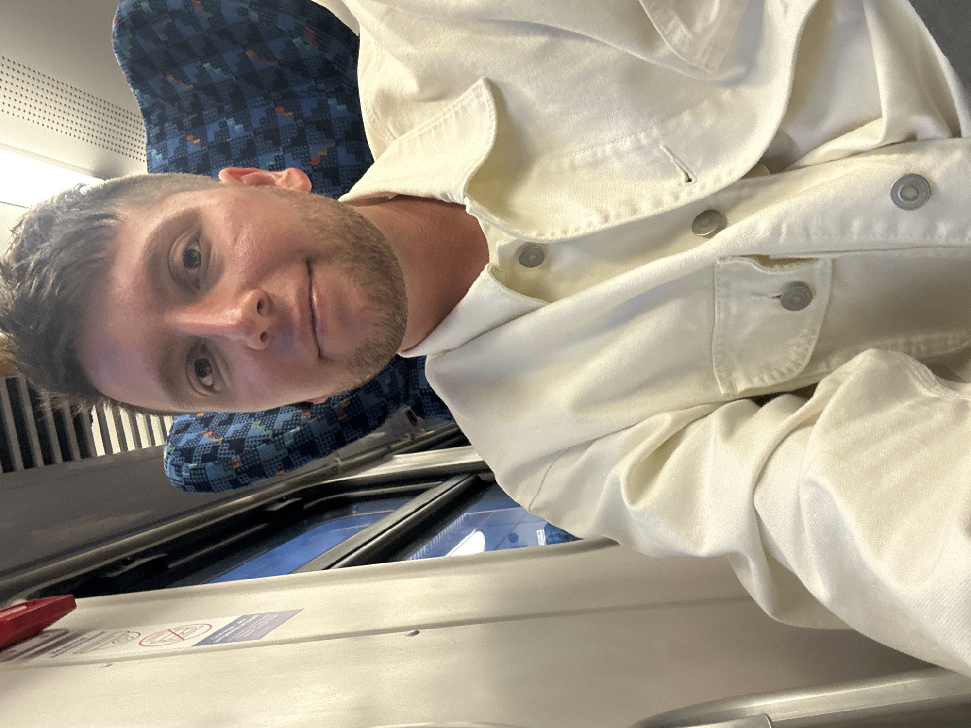 The image is a selfie taken on a train, showing a man wearing a white jacket. The interior of the train is visible, with a blue patterned seat behind the man. The window is partially visible, with the train's interior trim and a safety instruction label appearing in the foreground. The location is unknown, as the city of Balabanovo, Russia is not discernable from the visual elements present in the image. There is no verifiable evidence to support this location.