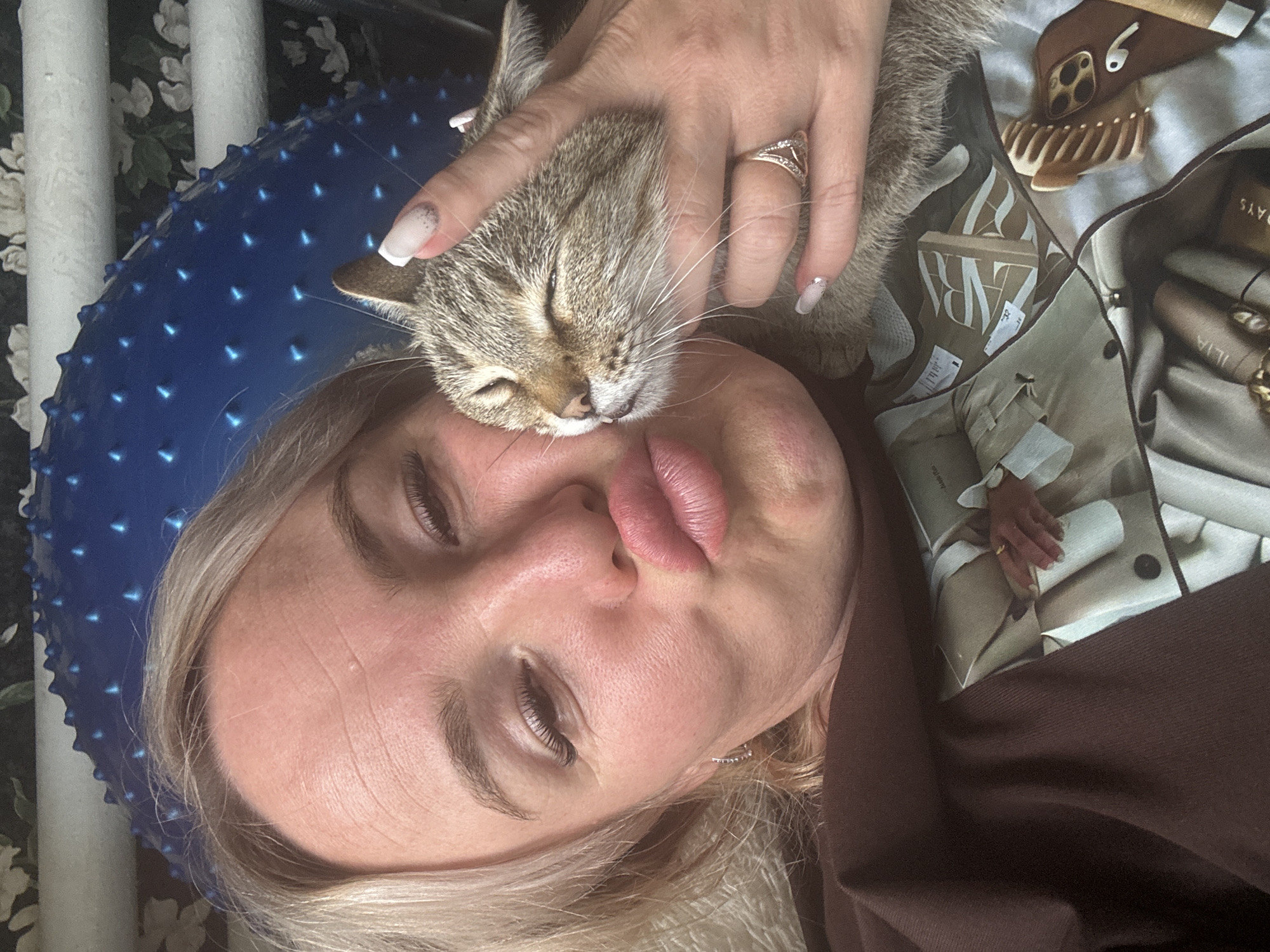 The media file shows a close-up, sideways selfie of a woman and a cat. The woman has blonde hair and is wearing makeup, including false eyelashes and lipstick. She is wearing a brown top. A tabby cat is nuzzling against her face. The woman's left hand is visible, resting on the cat's head. She is wearing a ring on her left ring finger.
The background includes a blue spiky massage ball and some decorative items. A patterned scarf is visible, featuring what appears to be a fashion-themed print with text that includes "ZARA" and "ILIA". There are also some cosmetic items like lipstick and mascara tubes scattered around. The overall impression is one of a casual, intimate moment between a person and their pet, possibly at home. The lighting suggests it is daytime. The image appears to be taken in Pozdyshki, Russia.
