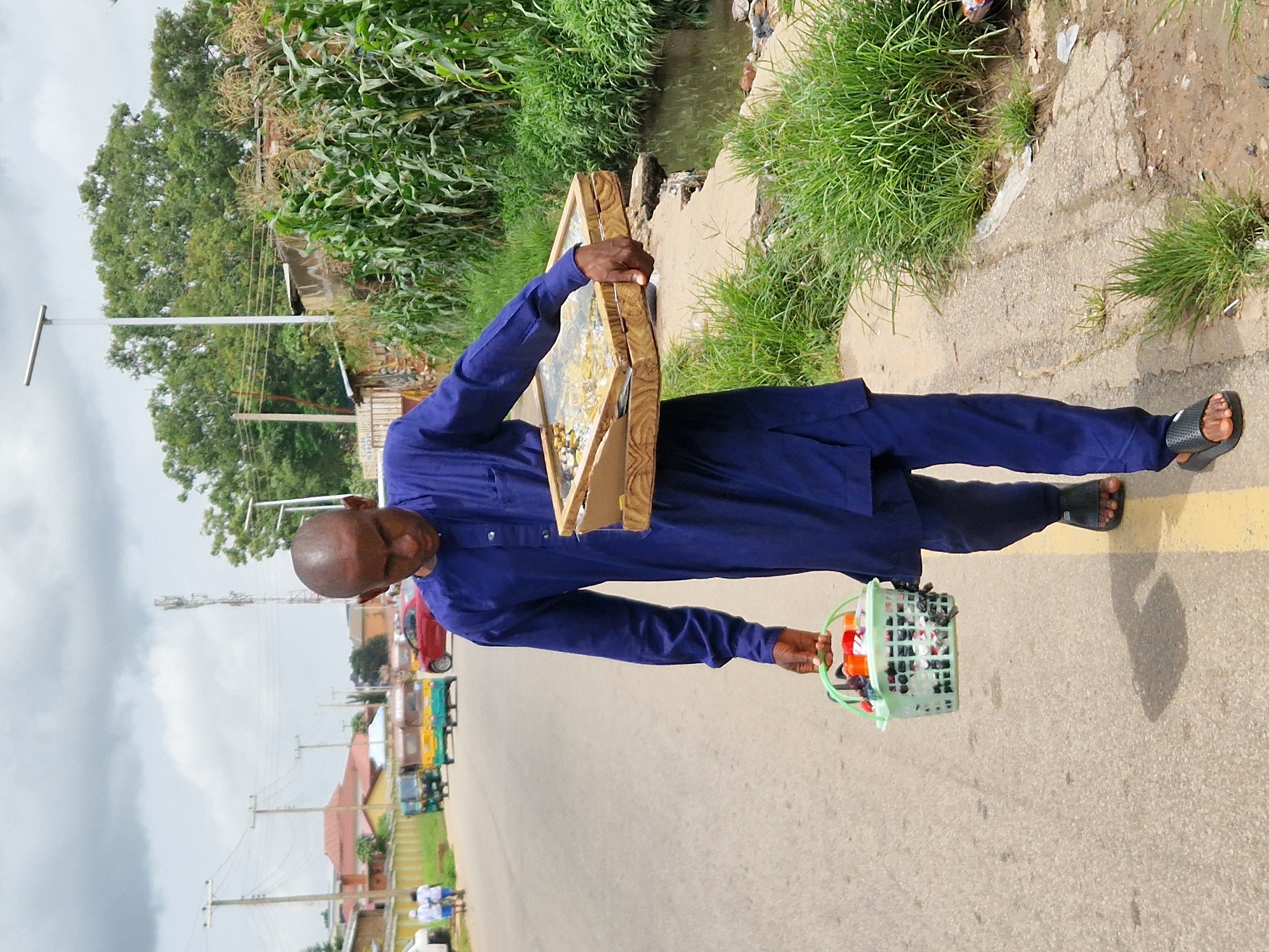 A bald man wearing a blue traditional outfit walks along a paved road in Jos, Nigeria. He is carrying a light green plastic basket filled with small items in his left hand and a wooden tray with a glass top containing what appears to be jewelry or trinkets in his right hand. The man is walking on the right side of the road, which has a yellow line marking. The environment includes a roadside drain with murky water and overgrown grass on the right. In the background, there are more trees, buildings, power lines, and a cellular tower. Vehicles, including a red car and several auto-rickshaws, are visible on the road in the distance. The sky is overcast with scattered clouds.
