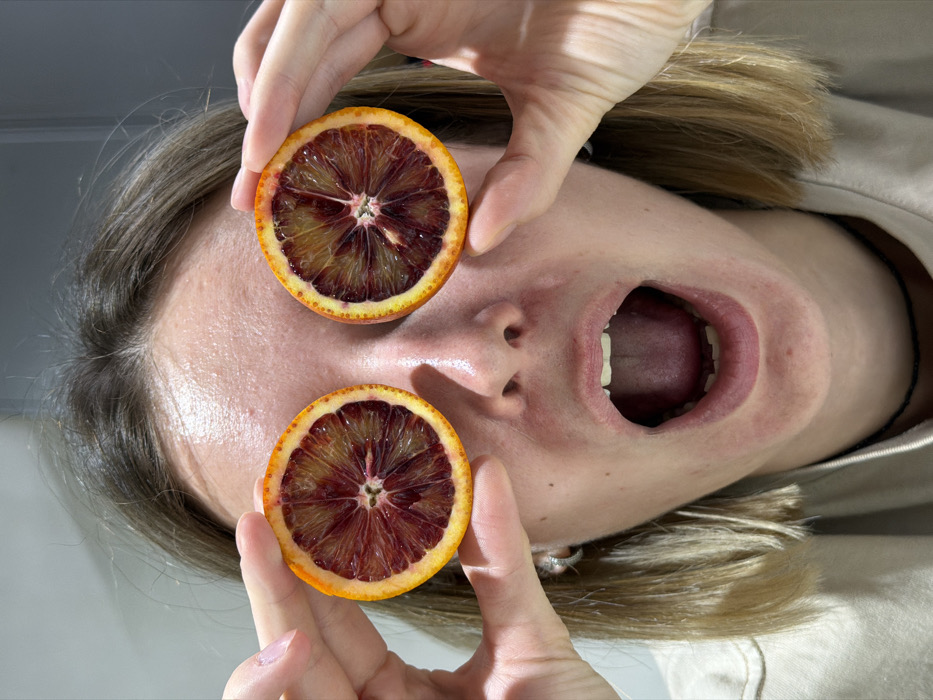 The image displays an adult individual in a frontal, close-up composition, situated in an indoor environment in Chaikovsky, Russia. The subject, possessing light brown, shoulder-length hair, is actively holding two horizontally sliced blood orange halves, one positioned over each eye, completely obstructing vision. Both hands are visible, with fingers manipulating the orange slices. The subject's mouth is wide open, displaying both upper and lower dental arches, indicating an exaggerated vocalization or expression of shock. Visible accessories include a thin black choker and a silver hoop earring in the subject's left ear. A beige or light-colored garment covers the lower portion of the subject's body within the frame. The background is an indistinct, uniformly light gray plane.