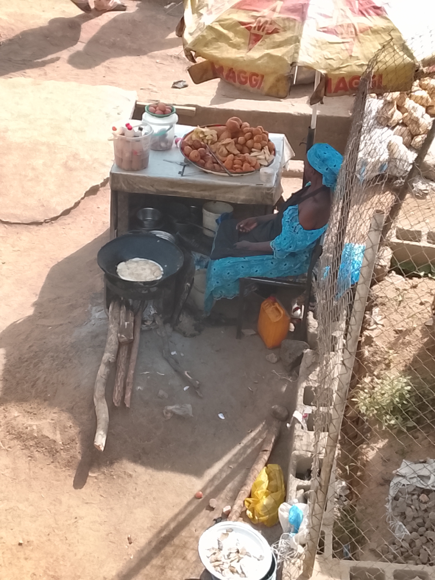 An outdoor food vendor operates from a makeshift stall in Gwong, Nigeria. A woman wearing a blue dress and head covering sits on a chair behind a small table laden with prepared food items, including fried goods. A cooking pot sits atop a wood-burning stove made of stacked stones and wood on the ground next to the table, with what appears to be dough or a flatbread inside. A Maggi brand umbrella provides shade over the stall. Nearby, a chain link fence encloses a dirt area. The ground is sandy and appears unpaved. A yellow container sits beneath the stall table. A white container rests on the ground near the fence.