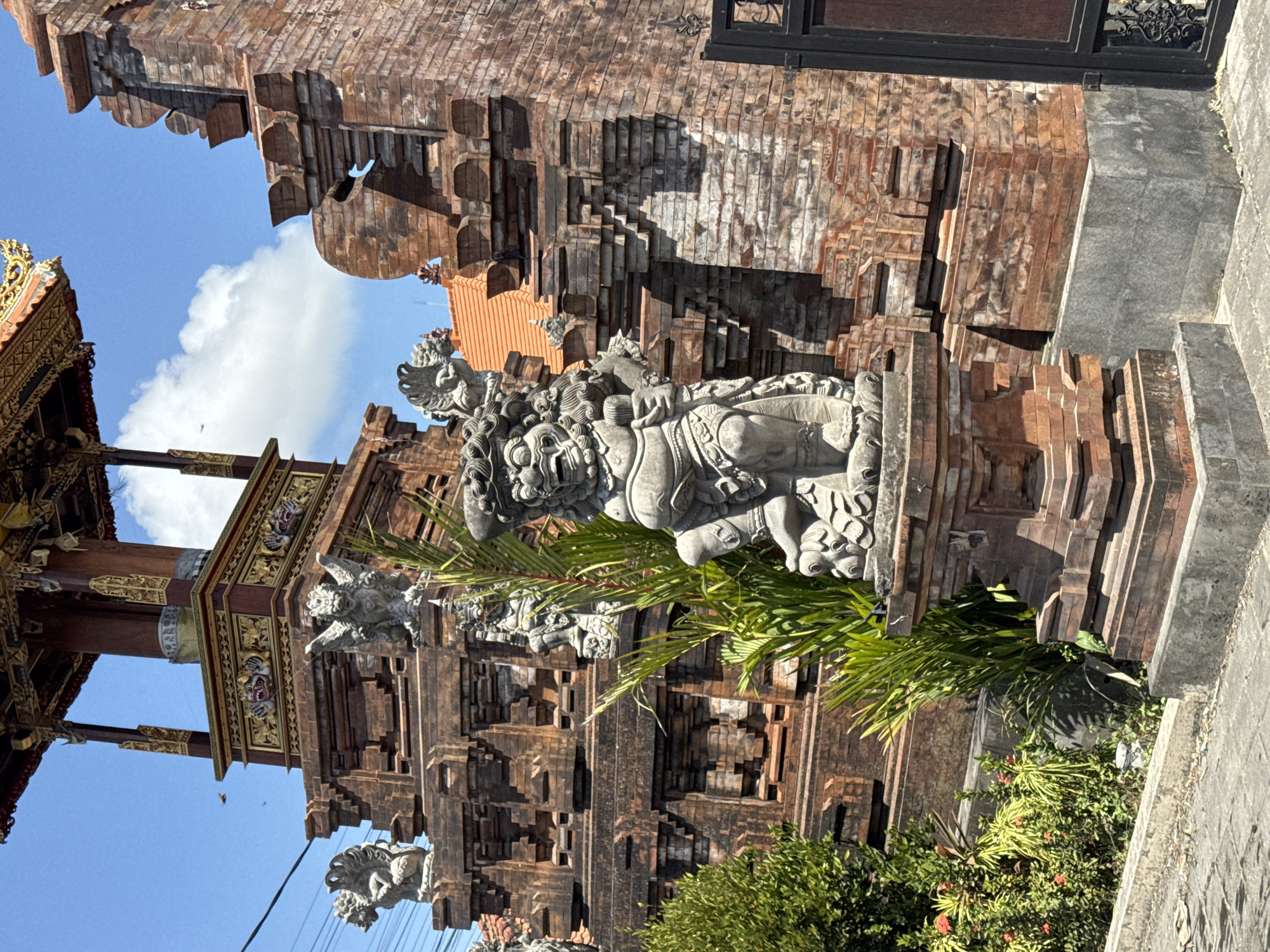 This image captures a detailed view of a traditional Balinese temple entrance structure, likely located in Denpasar, Indonesia, given the architectural style and common placement of such structures in the region. The composition is dominated by an ornate, multi-tiered wooden and brick edifice, adorned with intricate carvings and decorative elements, particularly on its upper levels. Two stone guardian statues, known as "Dwarapala," stand prominently in the foreground and mid-ground, depicted in fierce, dynamic poses, a common feature at temple entrances to ward off evil spirits. The main structure is constructed from weathered red bricks, exhibiting a stepped, layered design characteristic of Balinese temple architecture. Lush green foliage, including palm fronds and flowering bushes, is visible to the left and around the statues, softening the ruggedness of the stonework and brick. The background reveals a bright blue sky with scattered white clouds, indicating a clear, sunny day. The street in front of the temple appears to be paved with cobblestones. The overall impression is one of ancient artistry and spiritual significance, set within a tropical environment.