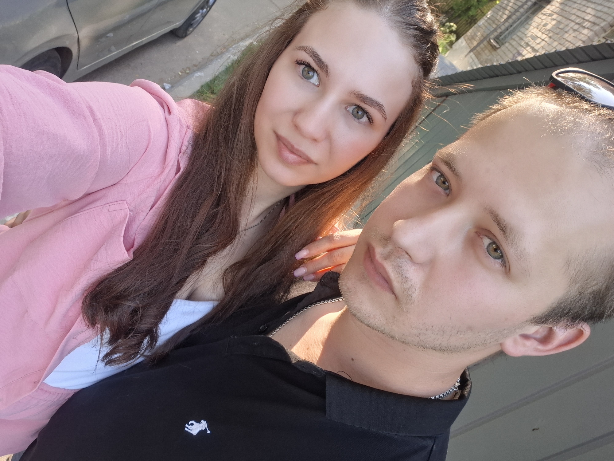 Two individuals, a woman and a man, are in the frame. The woman is wearing a pink garment over a white top, and the man is in a black polo shirt with a white emblem on the chest. The woman has her hand on the man's shoulder. The man is wearing a silver chain around his neck, and sunglasses are perched atop his head. Part of a vehicle is visible in the background, along with foliage and a structure with a slanted roof. The image appears to be taken outdoors in daylight. The location is Chaikovsky, Russia.
