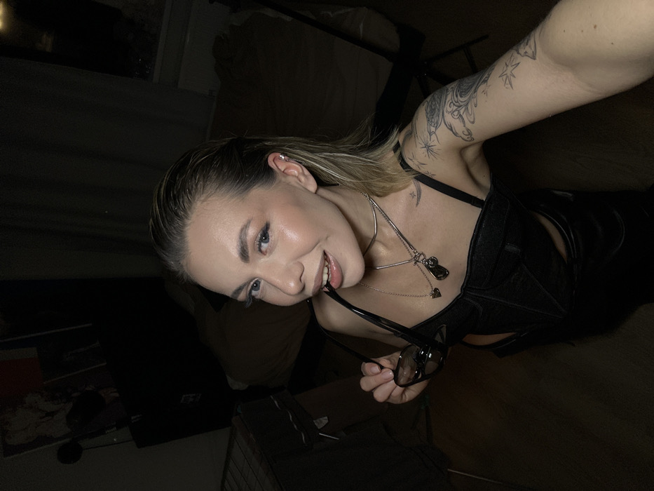 A young adult female is depicted in a dimly lit indoor setting. She has wet-look blonde hair, light skin, and is looking directly at the camera with a slight smile. She is wearing a black strappy top or bodysuit, multiple necklaces (including a choker, a chain with a heart and dog tag-like pendant, and another chain with a dark irregular pendant), and has an ear piercing on her visible left ear. Her left arm displays tattoos featuring celestial elements and abstract designs. She holds a pair of black sunglasses in her right hand, with one arm of the glasses positioned between her lips. The background is dark, revealing a clothes drying rack on the left, dark furniture behind her, and a window in the upper right showing a dark, unidentifiable exterior consistent with nighttime. Another dark object, possibly a chair, is visible on the right. The flooring appears to be dark wood or laminate. The precise location within Khimki, Russia, cannot be determined from the visual evidence presented in the image.