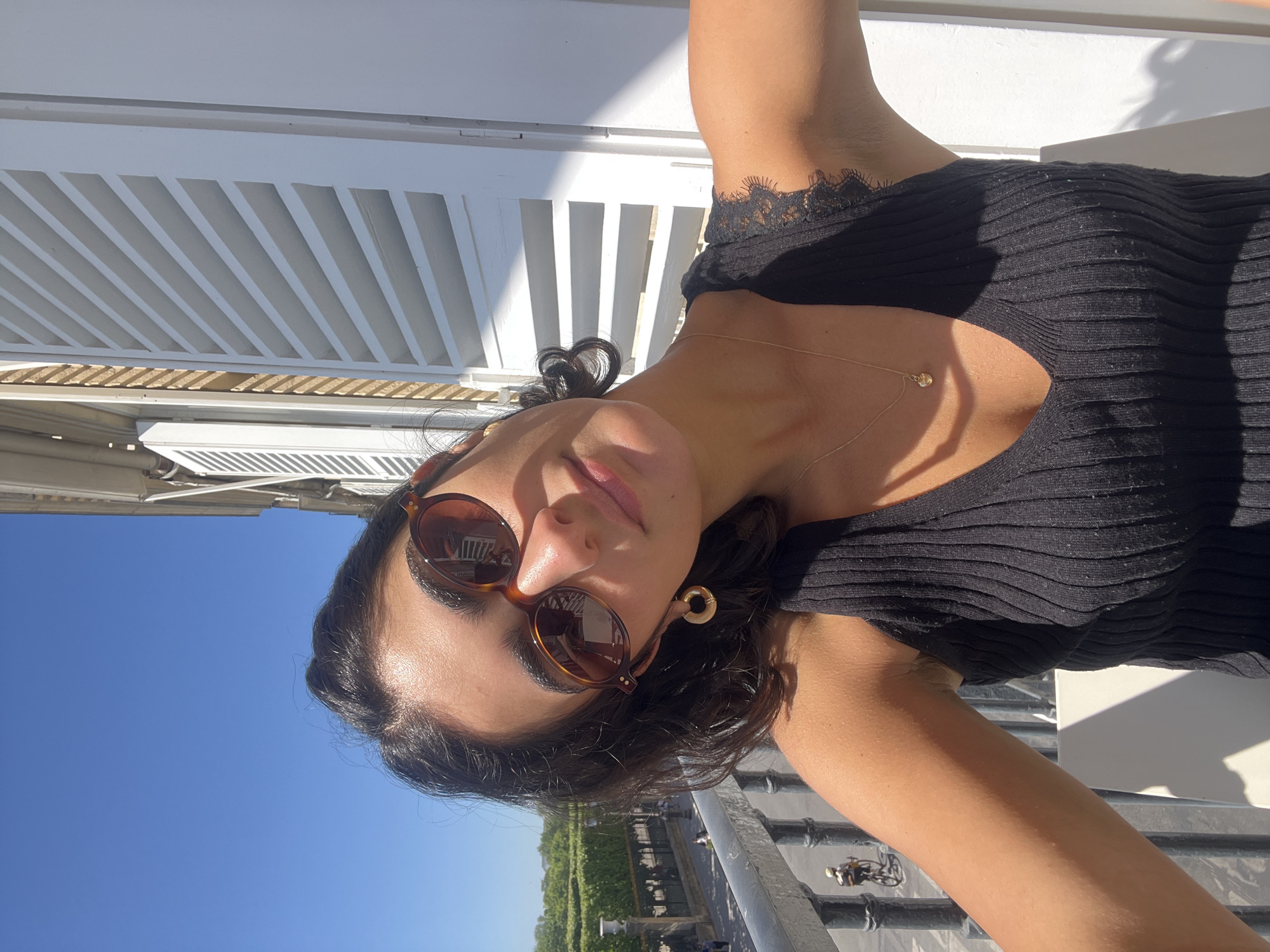 The image, originally rotated 90 degrees clockwise, captures a young woman taking a selfie outdoors on a bright, sunny day, likely on a balcony or terrace in Paris, France.
The primary subject is a woman with dark, wavy hair, wearing stylish brown, oval-shaped sunglasses. Her face, seen from a slightly upward angle, has a relaxed expression with a subtle smile. She is dressed in a black, ribbed top featuring a V-neck and delicate lace detailing along the neckline. Her accessories include a thin gold chain necklace with a small heart-shaped pendant and a prominent gold hoop earring on her visible right ear. She is holding the camera to capture the shot, with her arms slightly raised, creating a dynamic pose.
The setting behind her features bright white architectural elements, possibly louvered shutters or a pergola, which cast sharp, diagonal shadows, indicating strong, direct sunlight. Above and to the left (in the reoriented view), there is a clear, vibrant blue sky. Below her, a dark metal railing defines the edge of the balcony or terrace. Beyond the railing, a picturesque view unfolds, revealing distant green trees or gardens, suggestive of a park or boulevard. Faintly visible below are small figures of people and a cyclist, hinting at activity in the street or area beyond.
The strong sunlight, clear sky, and relaxed ambiance suggest it is daytime, likely mid-morning or afternoon, on a warm, pleasant day. The architectural style and distant greenery are consistent with the location context of Paris. No visible text appears in the image.