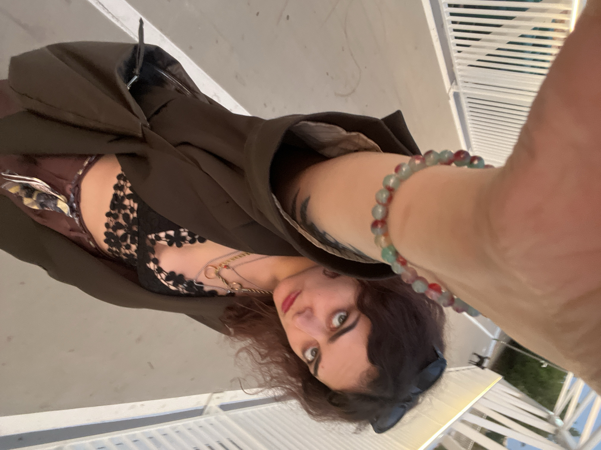 The image depicts a selfie-style, low-angle shot of an individual on a modern pedestrian bridge or elevated walkway, identified as being in Alcàsser, Spain. The subject, a person with wavy, reddish-brown hair, wears sunglasses pushed onto their head, a dark brown open jacket, and a black lace bralette. Multiple metallic necklaces with various pendants are visible around the neck, and a multi-colored beaded bracelet (red, green, light blue) is prominent on the right wrist, which is extended towards the camera. A dark tattoo is partially visible on the subject's inner right arm. The subject's gaze is directed towards the camera, and red lipstick is visible. The background features a light-colored paved walkway flanked by white railings constructed with vertical slats. In the distant background on the right, a small, dark-colored dog is visible walking on the path, alongside a street lamp. Beyond the bridge structure, green foliage and other structures are discernable. The scene is illuminated by natural light, creating some visible shadows.