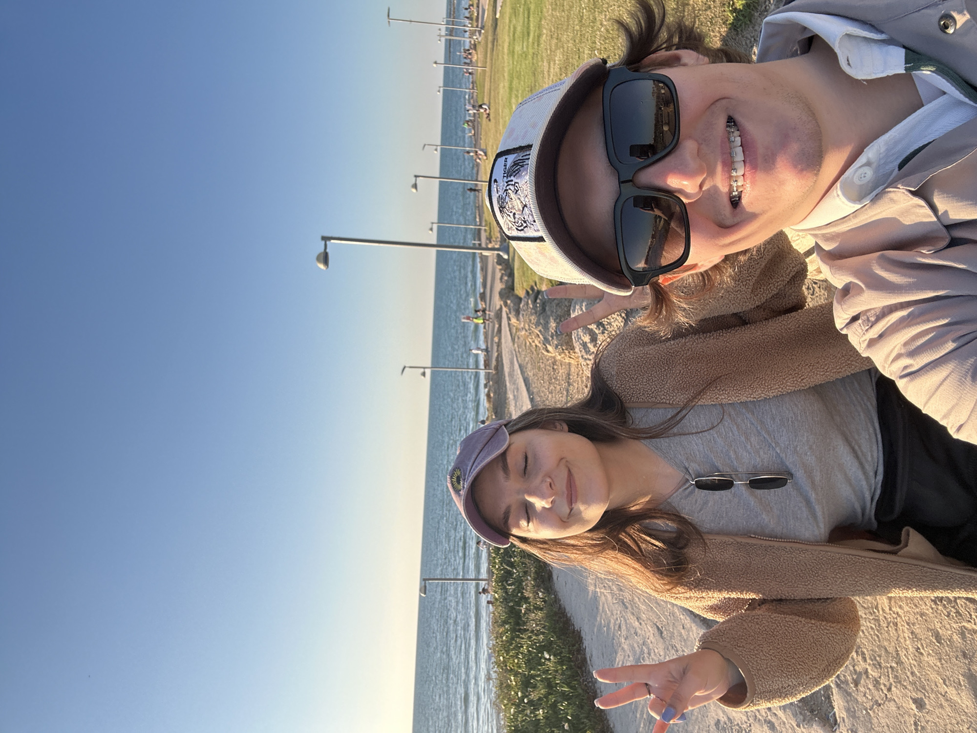 A selfie-style photograph, rotated 90 degrees clockwise, features two individuals – a man and a woman – posing outdoors by the sea, characteristic of a coastal promenade in Tel-Aviv, Israel. The setting sun suggests late afternoon or early evening.
In the foreground, on the right side of the rotated image, a man grins widely at the camera, showcasing braces on his upper teeth. He wears a light-colored baseball cap adorned with a black and white patch featuring an animal head, with the word "TIGER" subtly visible below. Dark, rectangular sunglasses obscure his eyes. He is dressed in a light-colored jacket or shirt layered over a white polo, and his left hand makes a peace sign.
Slightly behind and to his left, a woman smiles gently with her eyes closed or squinting. She wears a purple cap, a brown fleece jacket over a light grey t-shirt, and has dark sunglasses hanging from her neckline. Her right hand also displays a peace sign.
The background shows a calm body of water, likely the sea, extending horizontally across the image. A paved path or promenade runs alongside the water, with a strip of green grass visible further back. A line of distinct streetlights recedes into the distance along the water's edge. The sky above is clear and blue, gradually lightening towards the horizon on the left, indicating the low angle of the sun and contributing to the warm, golden hour lighting. Both subjects appear happy and relaxed, enjoying the coastal atmosphere.