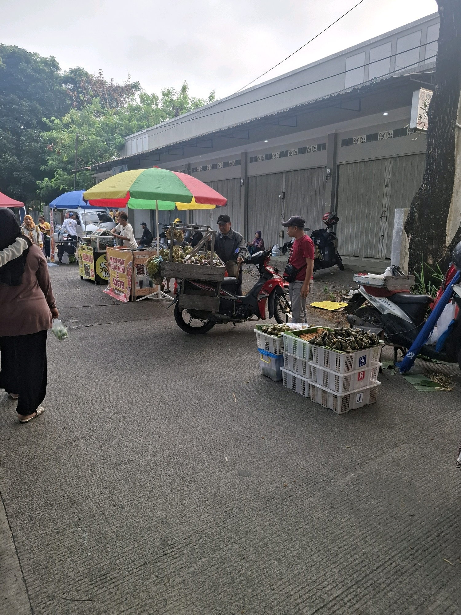 The image captures an outdoor scene, likely a street market, in the city of Karangtengah, Indonesia. The foreground is dominated by a paved road with a rough, textured surface. To the left, a woman wearing a brown headscarf and black clothing is walking away from the camera, carrying a transparent plastic bag with green items.
Further into the scene, several market stalls are set up. One stall, covered by a yellow and green umbrella, displays what appears to be durian fruit in a wooden cart attached to a red motorcycle. A person in a red shirt and a black cap is standing near this stall, looking to the left. Another market stall, under a blue umbrella, features a white vehicle and a sign that reads "MARTABAK TELUR MINI & OSTEL". People are visible behind this stall, suggesting a transaction or preparation of food.
In the background, a building with a corrugated metal roof and grey roller doors stretches across the frame. Trees with dense green foliage are visible on the left side of the image. Several motorcycles are parked around the market stalls, indicating transportation for vendors or customers. The sky is overcast with a soft, diffused light. The overall atmosphere suggests a bustling local marketplace.