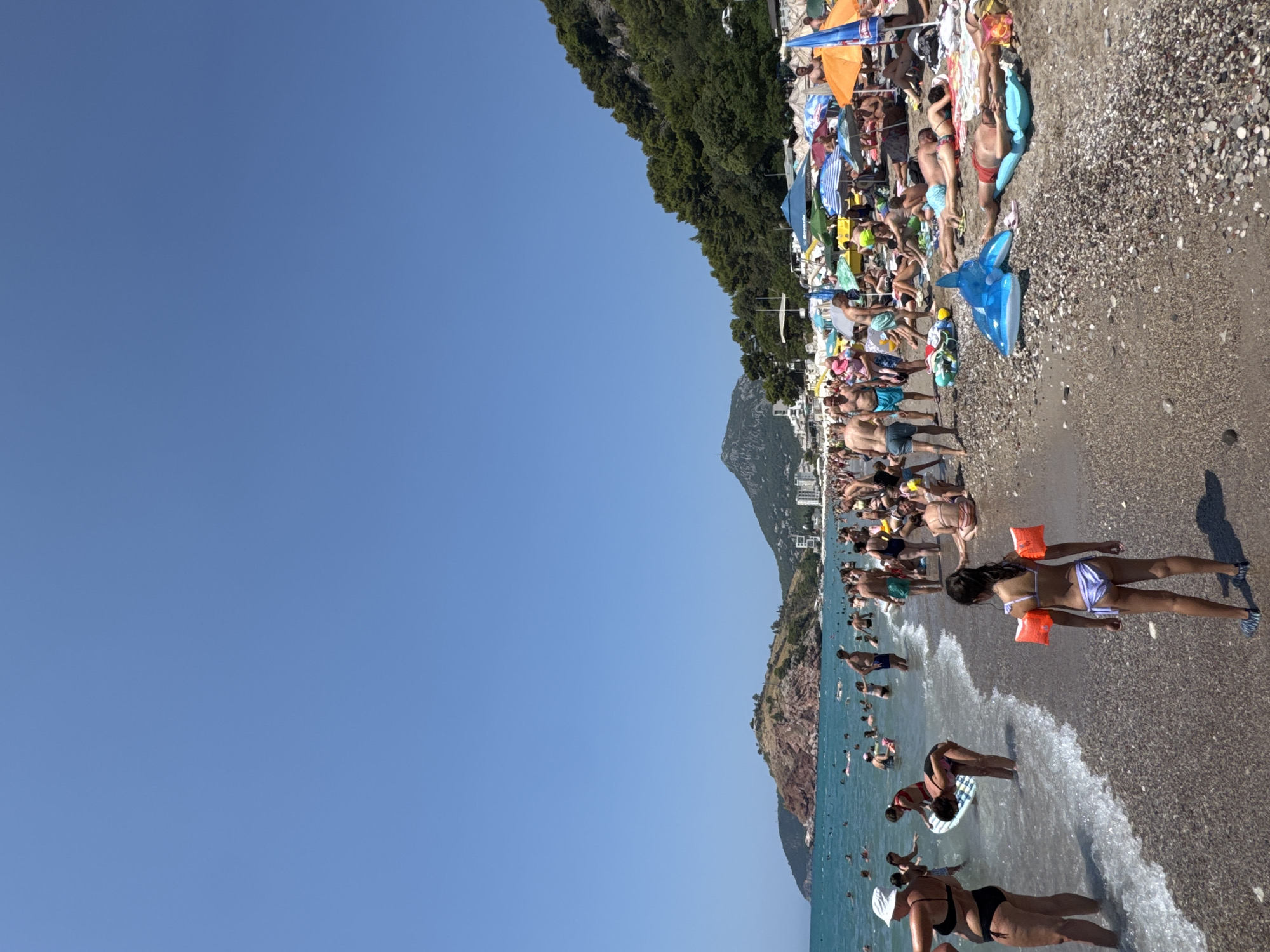 A crowded beach scene unfolds under a clear blue sky in Sutomore, Montenegro. The shoreline is populated with numerous people engaged in various beach activities. Several individuals are in the shallow water, while others are wading or swimming further out. A substantial number of people are relaxing on the pebble beach, some under colorful umbrellas, others basking in the sun. In the foreground, a young girl in a bikini and arm floaties stands facing away from the camera, looking towards the water. A blue inflatable dolphin toy lies on the pebbles nearby. In the midground, beachgoers are spread across the sand and water. Behind the beach, a verdant, tree-covered mountainside rises steeply, with buildings and more people visible along its base. A prominent rocky hill with some structures on its summit is situated in the distance on the left, bordering the sea. The overall atmosphere suggests a lively and popular summer day at the seaside.