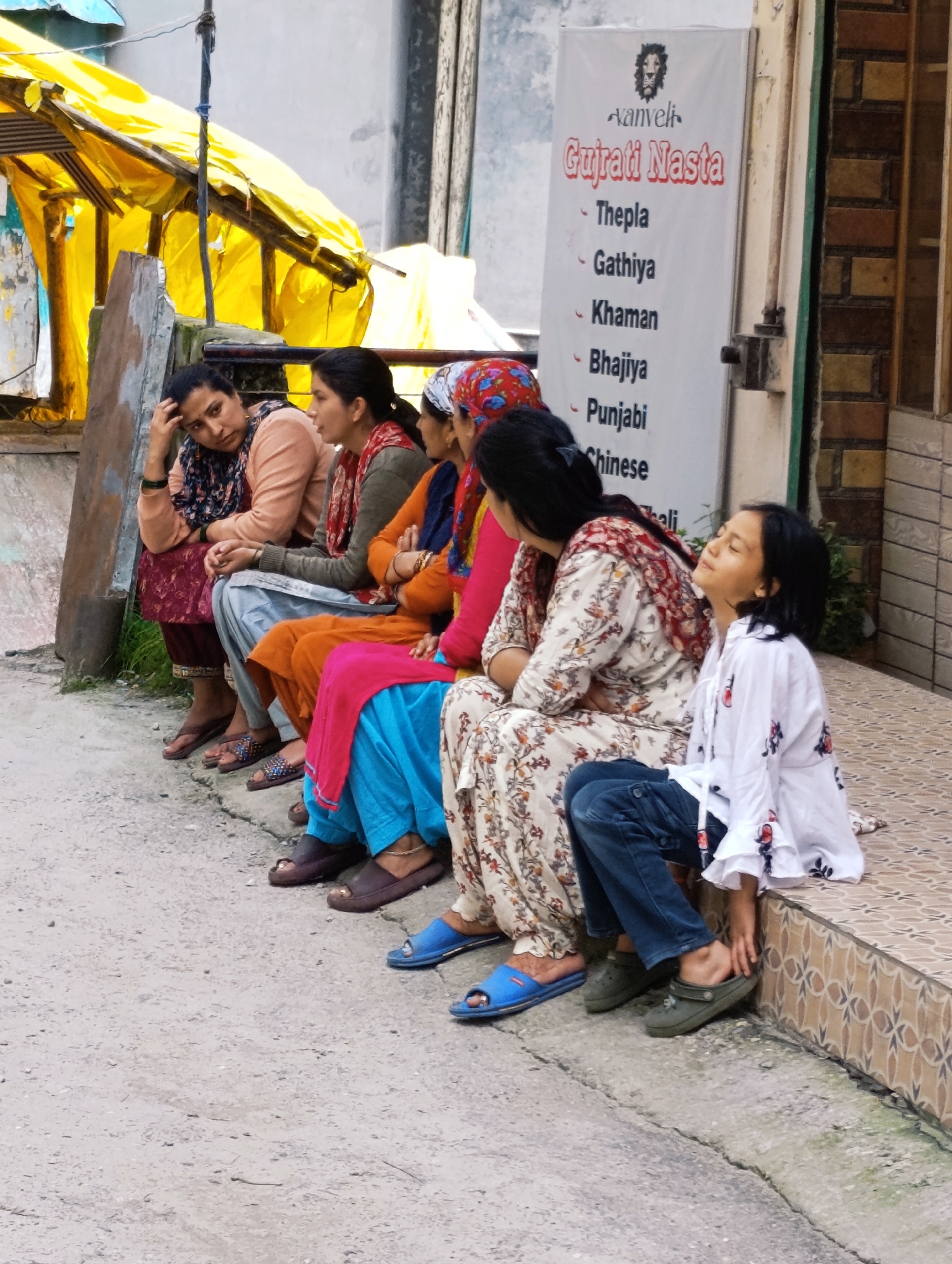 A group of five individuals, appearing to be women and a young girl, are seated on a paved surface outside a building in Vashisht, India. They are arranged in a line from left to right. The woman on the far left, wearing a light peach-colored top and a patterned headscarf and skirt, has her hand on her forehead. To her right, a woman in an orange and red outfit with a patterned scarf is speaking. Next to her, a woman in a bright orange kurta and blue salwar is seated. Following her, a woman in a cream-colored floral print kurta and matching salwar is looking down. On the far right, a young girl wearing a white top with floral accents and blue jeans is seated, looking upwards with her eyes closed. Behind them, a white banner with text, including "Aanveli Gujarati Nasta" and a list of food items like "Thepla," "Gathiya," and "Punjabi," is affixed to the building. A yellow awning is visible to the left of the group. The environment suggests a casual outdoor seating area.