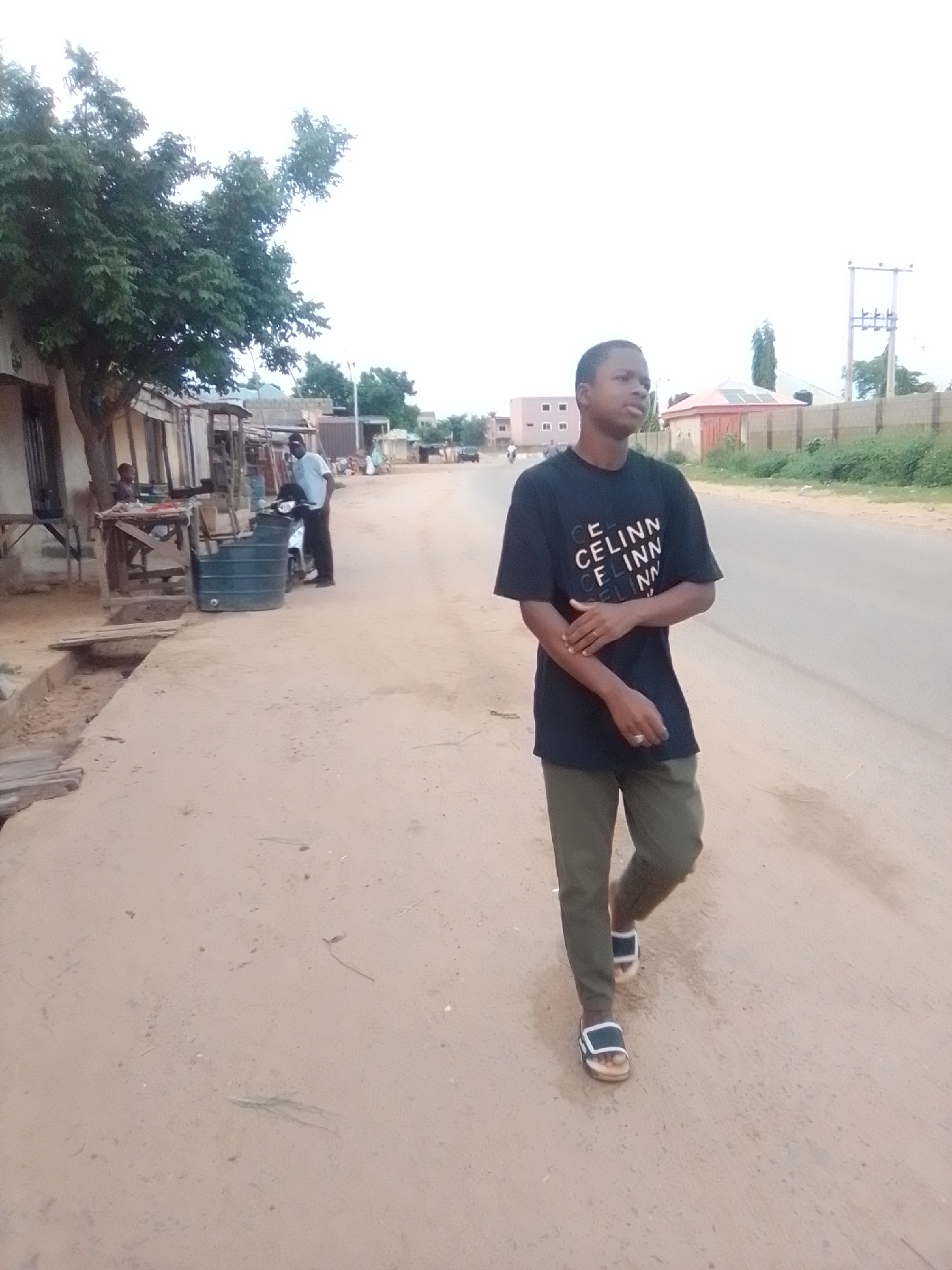 The image depicts a street scene in the city of Galadima, Nigeria. A young man wearing a black t-shirt with white lettering and green trousers is walking towards the right of the frame on a dirt road. He is wearing sandals. To the left of the road, there are several market stalls and buildings constructed with wood and corrugated metal. A large dark green water tank is visible near one of the stalls. A man in a white shirt and dark trousers is standing next to a motorcycle near the stalls. Further down the road, there are more buildings and trees. The sky is overcast and appears bright.